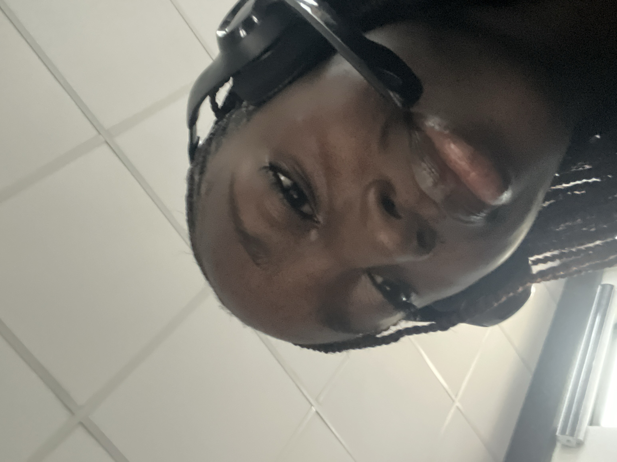 A close-up, upward-facing shot shows a Black woman with braided hair wearing black headphones. Her eye is visible, as is her nose and mouth. The background is a white, tiled ceiling. A window frame is partially visible in the lower right corner. The image provides no definitive details to confirm a location in Karum, Nigeria.