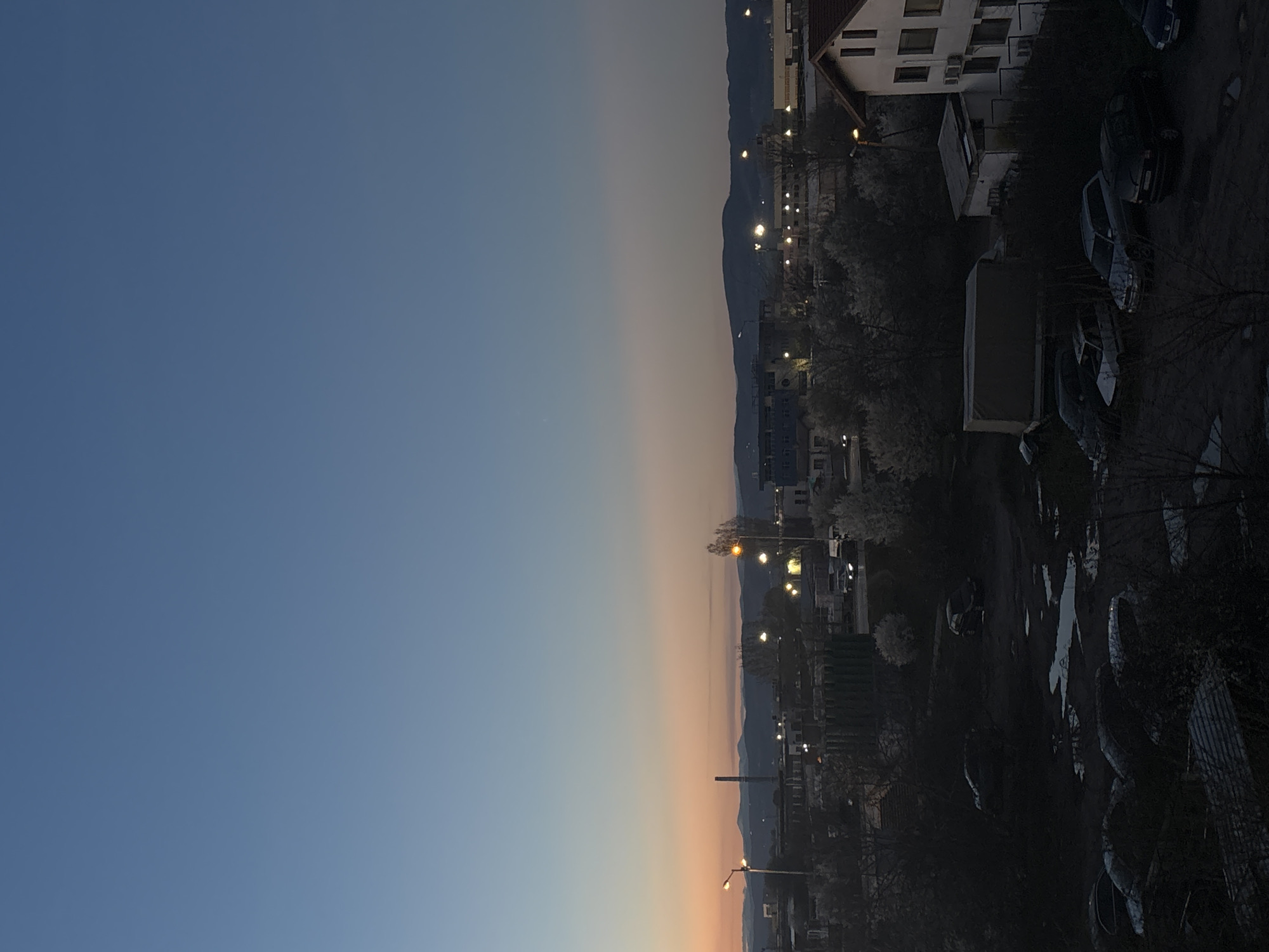 The image shows a cityscape at dusk, likely in Sofia, Bulgaria. The left side of the frame is dominated by a gradient of blue sky, transitioning to orange near the horizon. The right side of the image shows a residential area, including apartment buildings, parked cars, and streetlights. Mountains are visible in the background. The scene is mostly dark, with artificial lighting illuminating the buildings and streets.