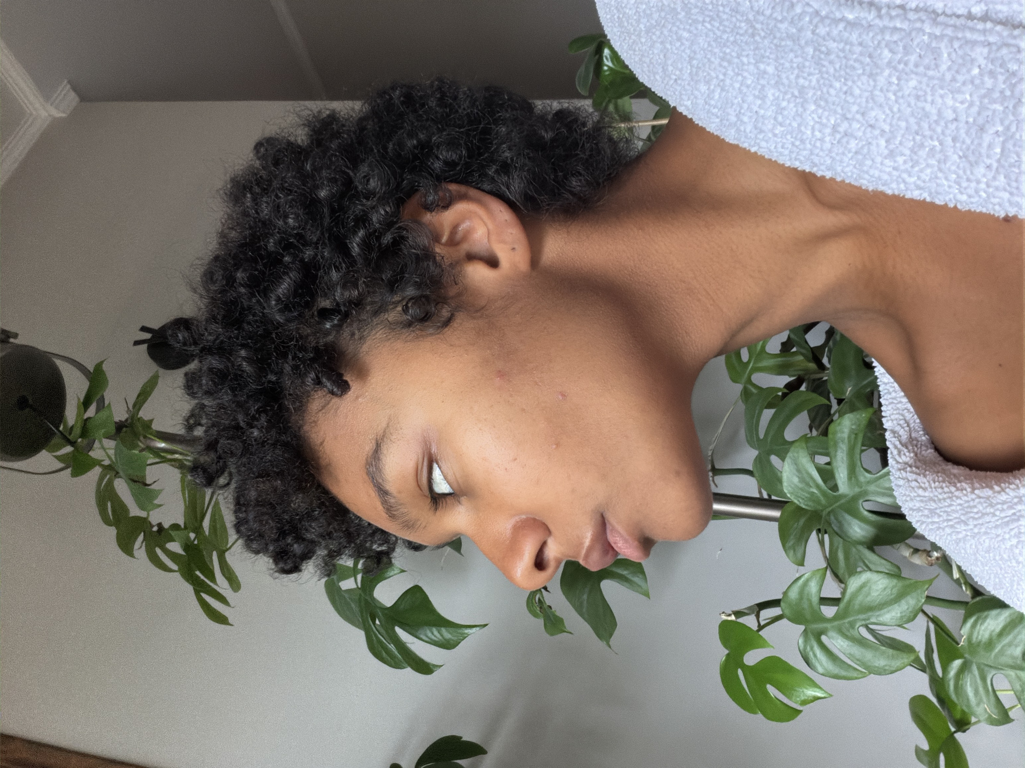 An eye-level shot captures a young adult with dark, curly hair, seen in a relaxed, contemplative pose. The subject's head is tilted slightly forward, with their gaze directed towards the upper right of the frame (from their perspective). Their skin tone is medium-dark, and their features are clear, with a neutral expression. A subtle glow highlights parts of their face and neck. They are partially covered by a white, textured towel or bathrobe draped over their right shoulder and upper torso, hinting at a post-shower or relaxed state.
The background consists of a plain, light-colored wall. Lush green plants with distinctive lobed leaves, resembling Rhaphidophora Tetrasperma or a similar vining species, frame the subject. One plant trails downwards from a dark, suspended pot in the upper left background, while another appears to grow vertically on a support pole, positioned behind the person's neck and shoulder area. The lighting is soft and natural, suggesting an indoor setting during the daytime. The overall ambiance is calm and serene. No text is visible in the image. The scene is set in Champs-sur-Marne, France.