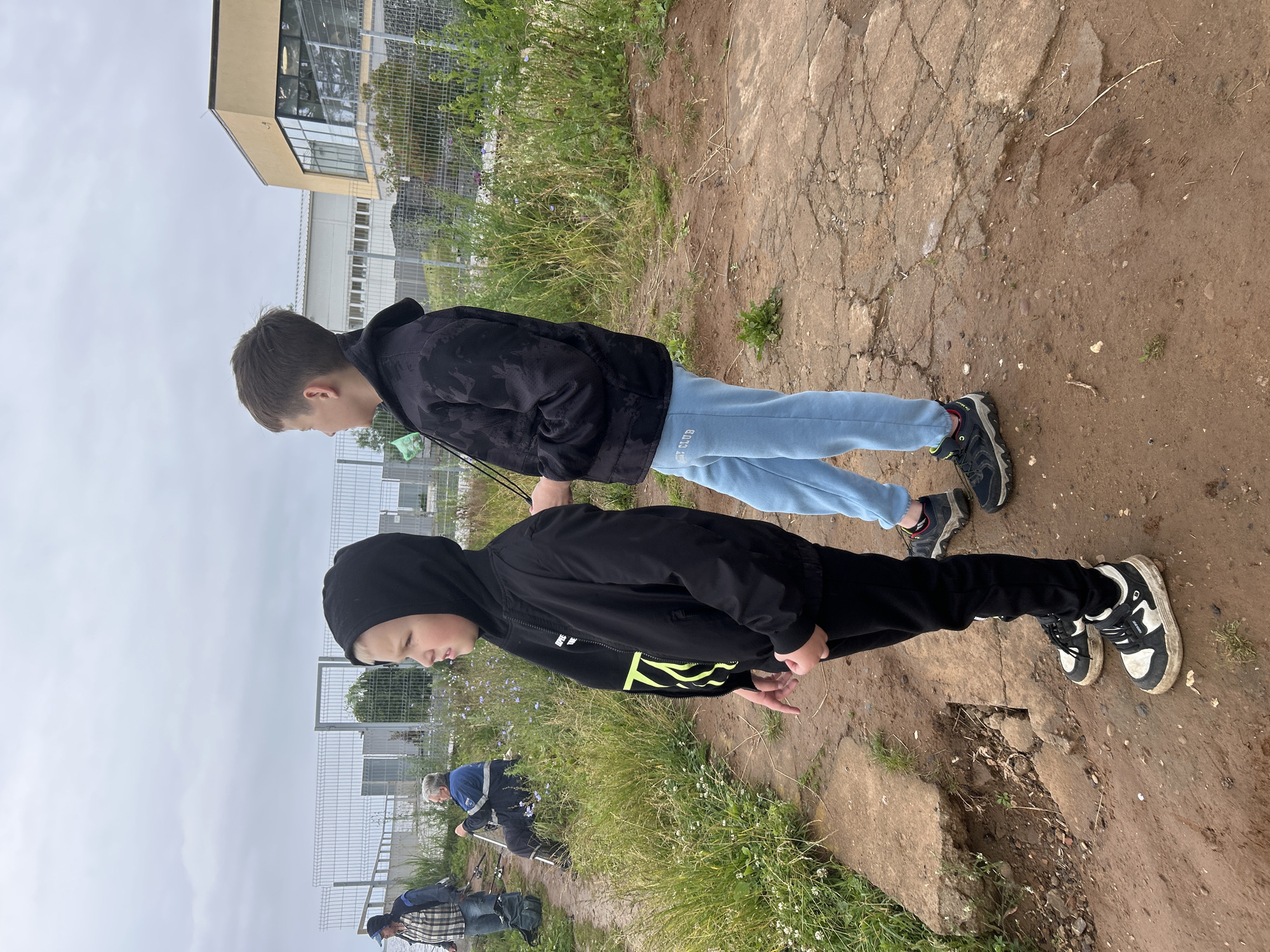 Two young boys are standing on a dirt path next to a grassy area. The boy in the foreground is wearing a black hooded jacket with a bright yellow graphic and black pants. He is looking down. The boy behind him is wearing a black patterned jacket and light blue sweatpants. He is looking to his left. In the background, a building with large windows and a fence can be seen. Further in the distance, other people are visible, some appearing to be fishing near a fence line. The ground is a mix of dirt and cracked concrete. The sky is overcast, suggesting it might be a cloudy or rainy day. The overall setting appears to be outdoors, possibly a park or recreational area. Text visible on the light blue sweatpants says "MY CLUB".