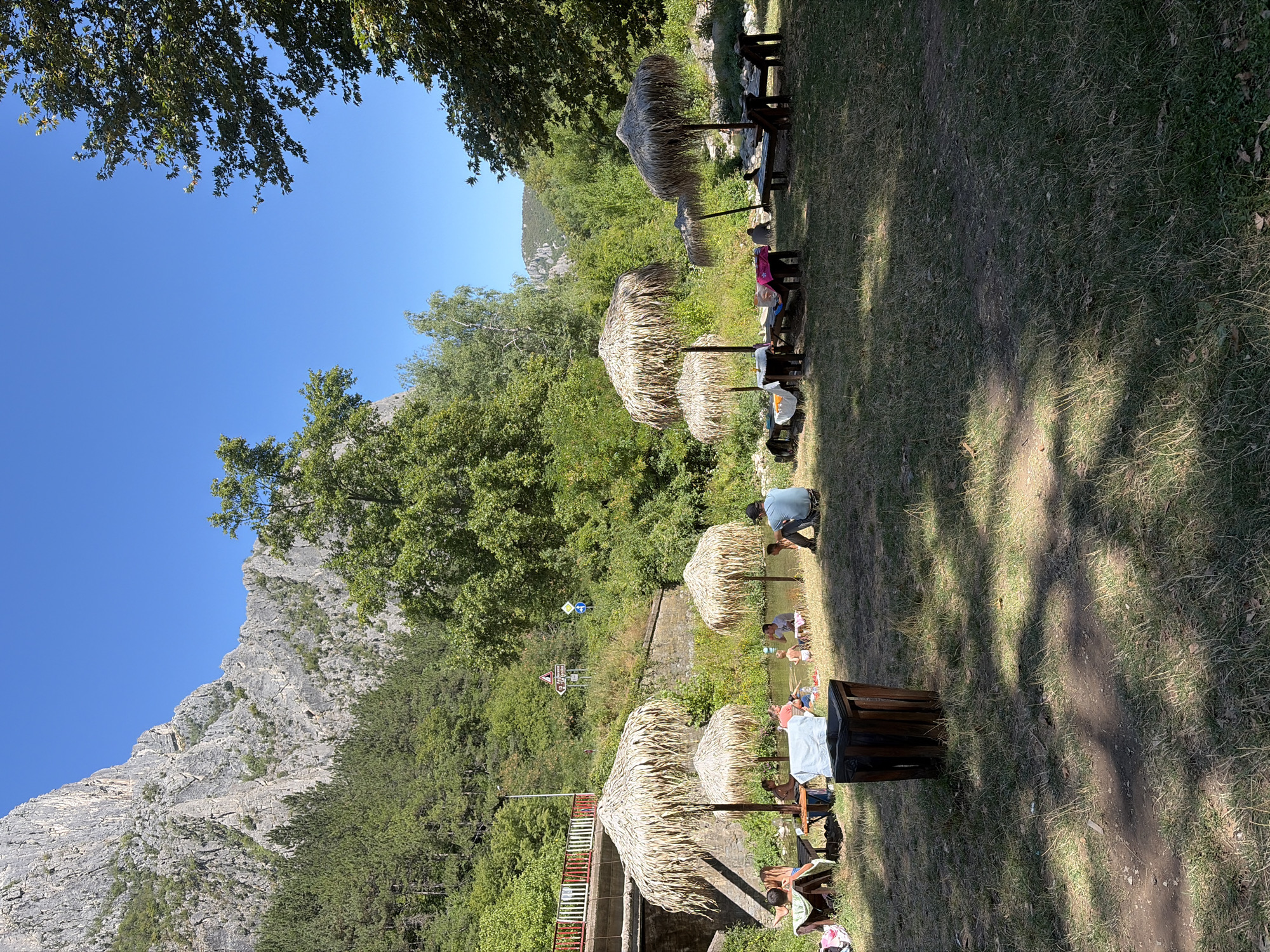 In Zgorigrad, Bulgaria, a tranquil scene unfolds by a river. Several thatched umbrellas provide shade for small groups of people relaxing on lounge chairs and near picnic tables. Some individuals are seated, while others appear to be wading in the shallow river water, which is surrounded by lush green trees and a rocky mountain face. The area is a designated relaxation spot, with a trash can visible in the foreground. The overall atmosphere suggests a peaceful summer day spent enjoying nature.