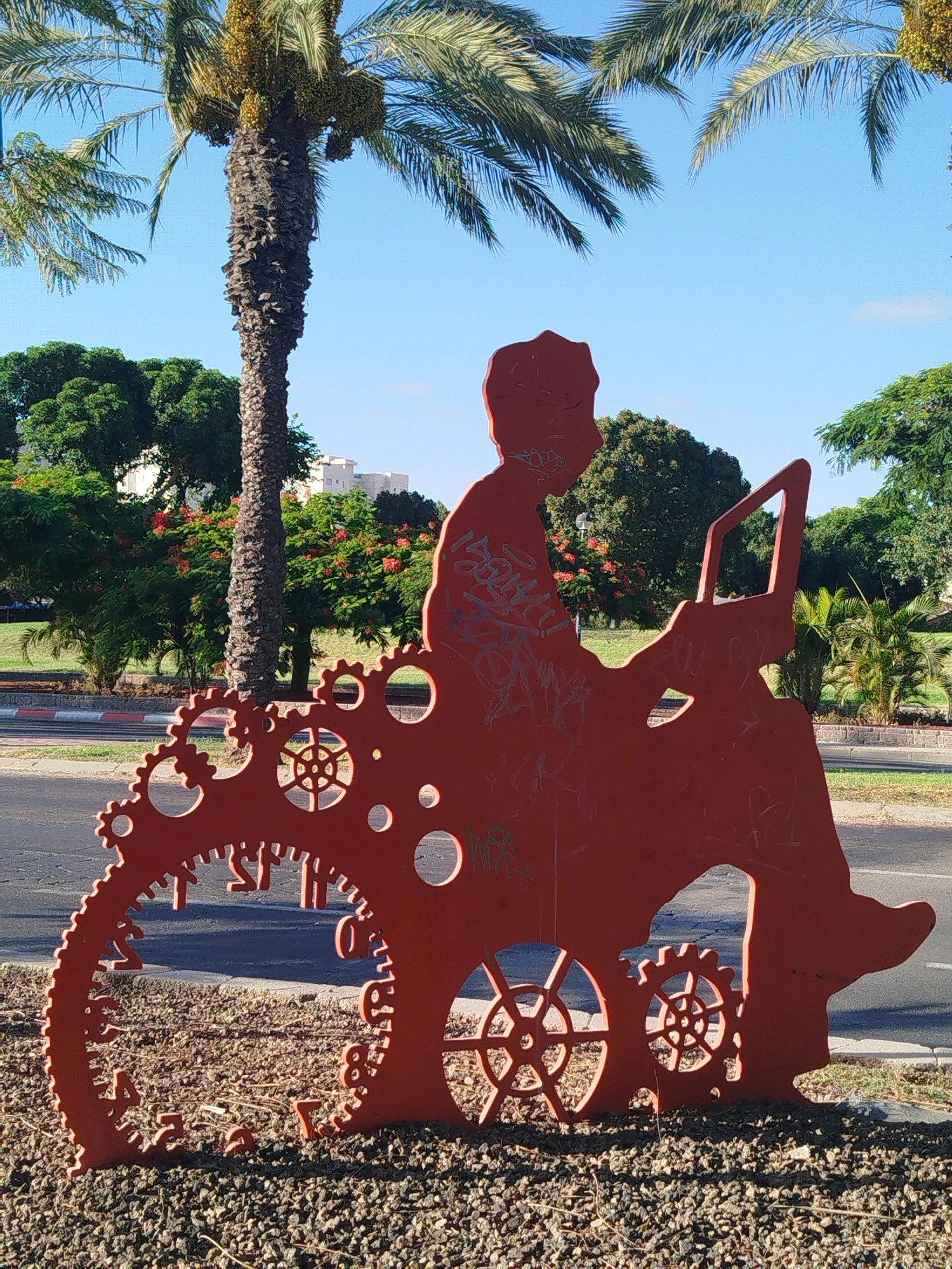 This is a full shot of a public art installation in Ashkelon, Israel. The sculpture, made of bright orange metal, depicts a person in silhouette seated with a laptop. The figure is integrated into a large gear mechanism that forms the base of the sculpture, resembling a bicycle wheel with gears. The sculpture is situated outdoors, in what appears to be a park or a roadside median, with gravel on the ground and a paved road visible in the background. Behind the sculpture, lush green trees, including palm trees, and flowering trees with red blossoms are visible. The sky is clear and blue. The sculpture shows some signs of graffiti, with red spray paint visible on the metal. In the distance, a few multi-story buildings can be seen, suggesting an urban or suburban setting.