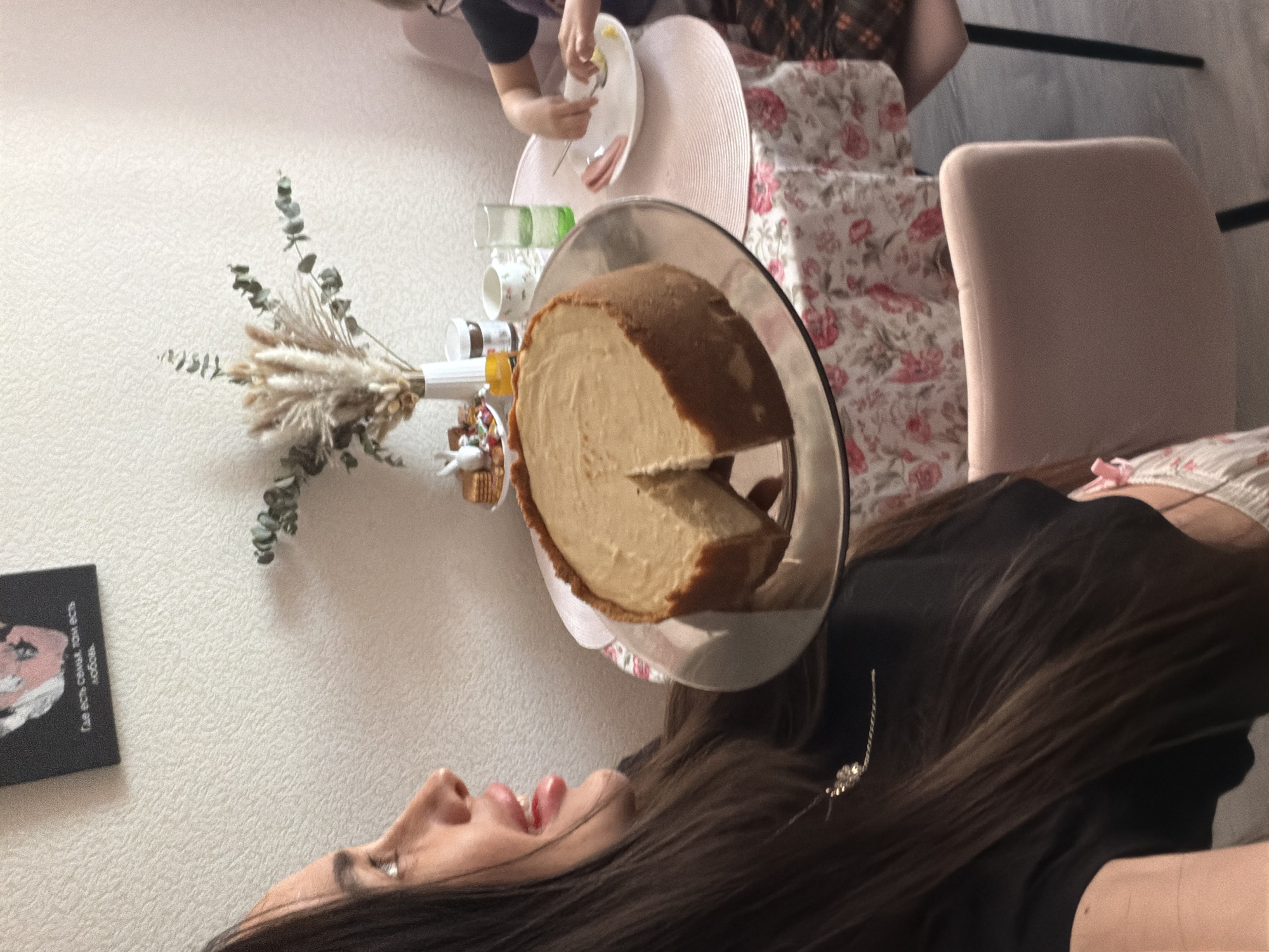 The image appears to be taken indoors, possibly in an apartment. A woman with long dark hair is visible in the foreground, partially obscured by a slice of cake on a glass plate. The cake is sitting on a small table. Other individuals are present, one eating food on a plate, and another sitting on a chair with a floral pattern. There is a vase with dried flowers on the table, along with some glasses and small decorative objects. A book with text in Russian sits on the wall. It is difficult to confirm if the location is indeed Saint Petersburg, Russia, without additional contextual cues in the image.