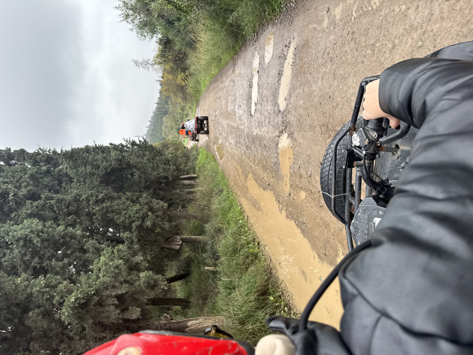 The image depicts a first-person perspective from an individual operating an all-terrain vehicle (ATV) on a muddy, unpaved road in Çiftlikköy, Turkey. The driver wears a red helmet, partially visible on the left, and a dark, possibly padded, jacket, with one hand gripping the right handlebar of the ATV. Ahead, two other ATVs are in a procession: the lead ATV is orange, followed by a maroon ATV. The rider of the maroon ATV is covered by a light-colored, possibly white, hooded garment. The dirt road is extensively marked with tire tracks and numerous large puddles of brown water, indicating recent moisture. To the left, a dense line of tall, slender, dark green coniferous trees borders the road, with an undergrowth of grass and smaller vegetation. The right side of the road features varied green and yellowish-brown foliage. In the distant background, a tree-covered hill is visible. The sky is uniformly overcast and light gray.