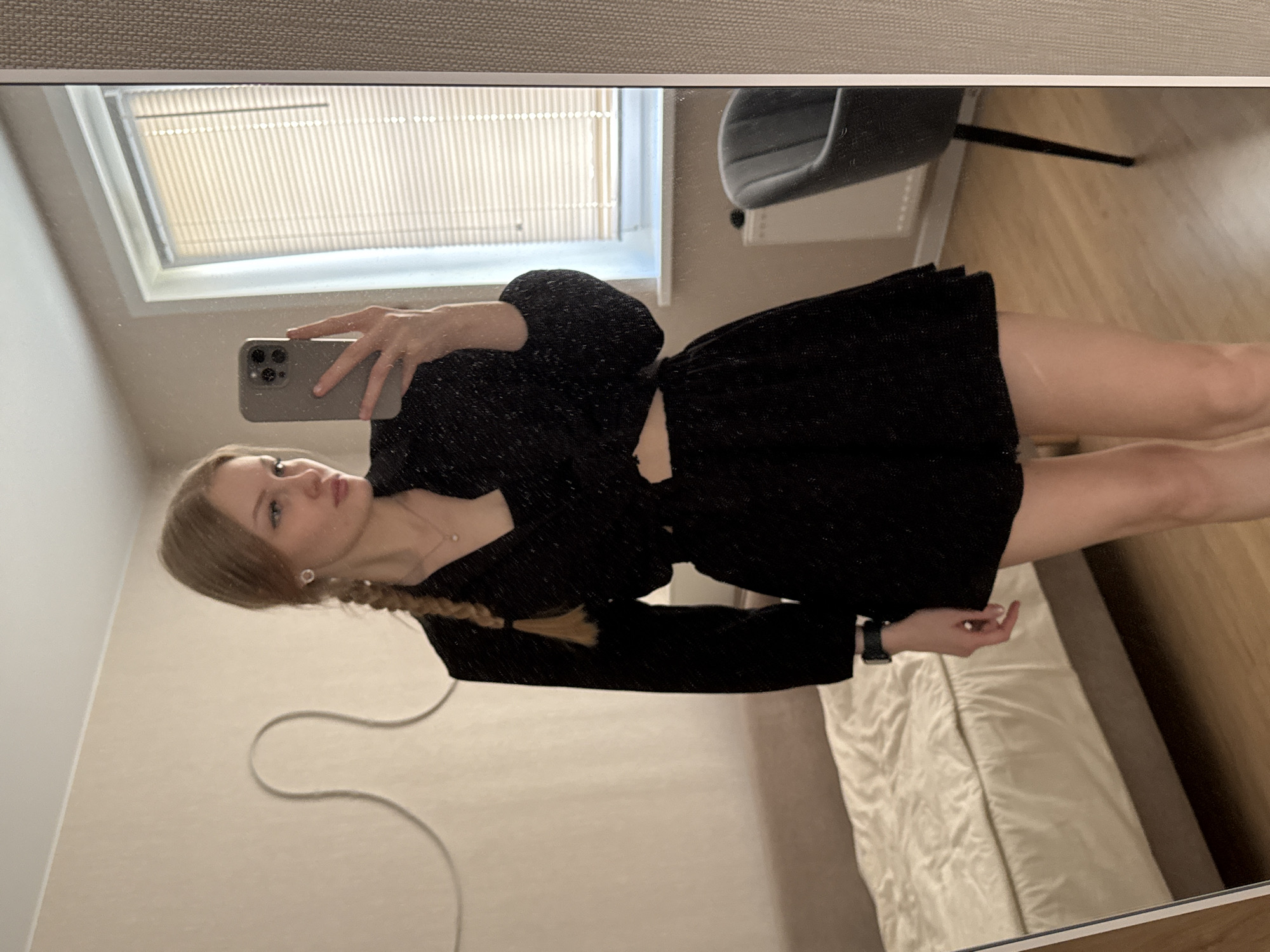 A young woman is posing in a mirror, likely taking a selfie. She is wearing a black, short dress or a two-piece outfit with a top and a skirt. She has blonde hair styled in a braid. The reflection in the mirror shows parts of a room: a bed with white bedding, a chair, and a window with blinds. It can be assumed that the location is a bedroom or a dressing room. The image was taken from the woman's perspective, looking at herself in the mirror. The location in the city of Perm, Russia is not determinable from the image.
