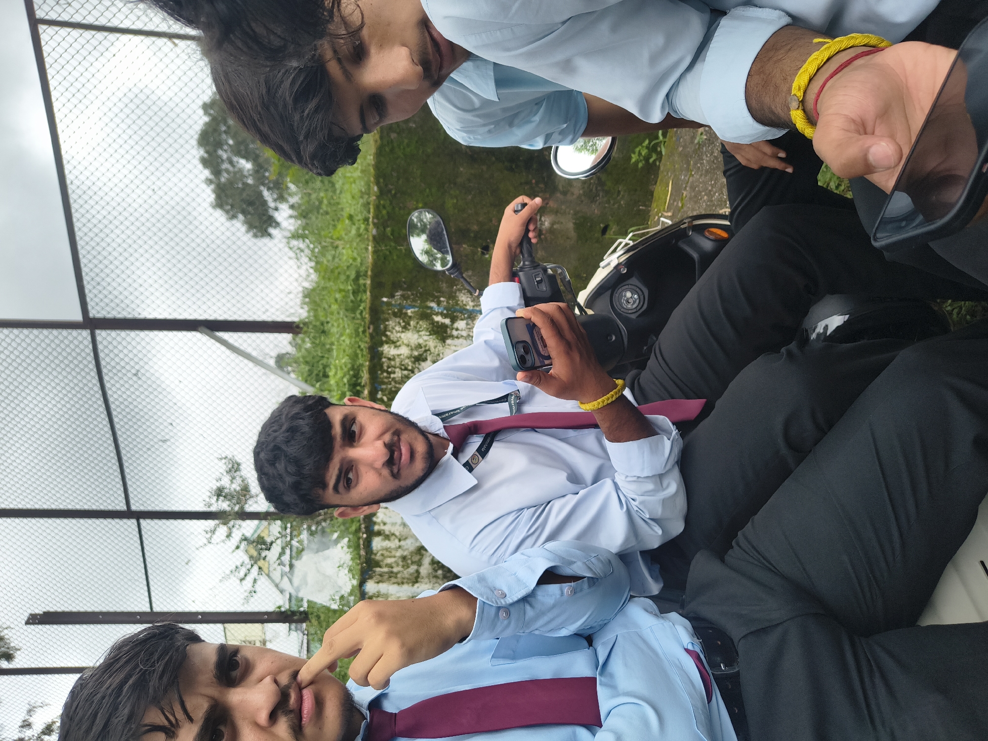 The image captures a group of young men in light blue collared shirts and dark trousers, possibly students, on or near a scooter. The setting appears to be outdoors, with a fence and greenery in the background, suggesting a location like a college campus or a park. One man in the foreground is holding a smartphone, while another is touching his lip. The presence of a scooter implies mobility or a recent journey. The overall atmosphere is casual and social. The overcast sky suggests it might be a cloudy day.