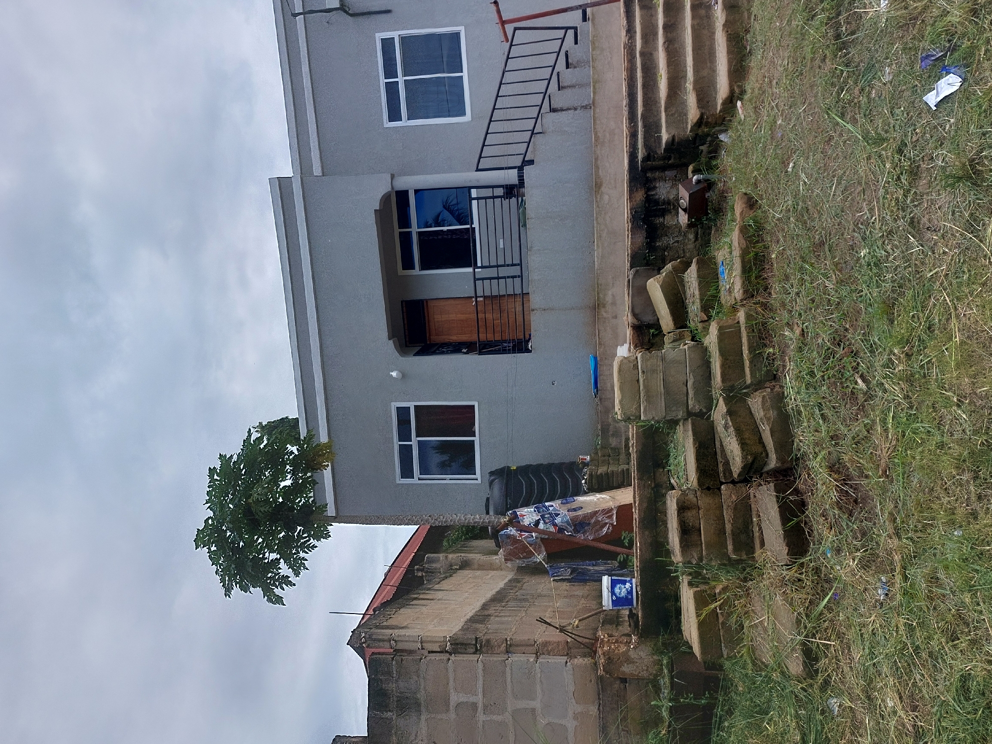 A two-story gray building with white-framed windows and a small balcony is the focal point of the scene. A stairway with a black railing leads to the second floor. The building is situated next to a grassy area with a few scattered pieces of trash. Adjacent to the building, a smaller structure with a red roof is visible, constructed of brick. A water tank and various objects covered with plastic are visible on the roof of the smaller building. The sky is overcast. The location is Dar es-Salaam, Tanzania.