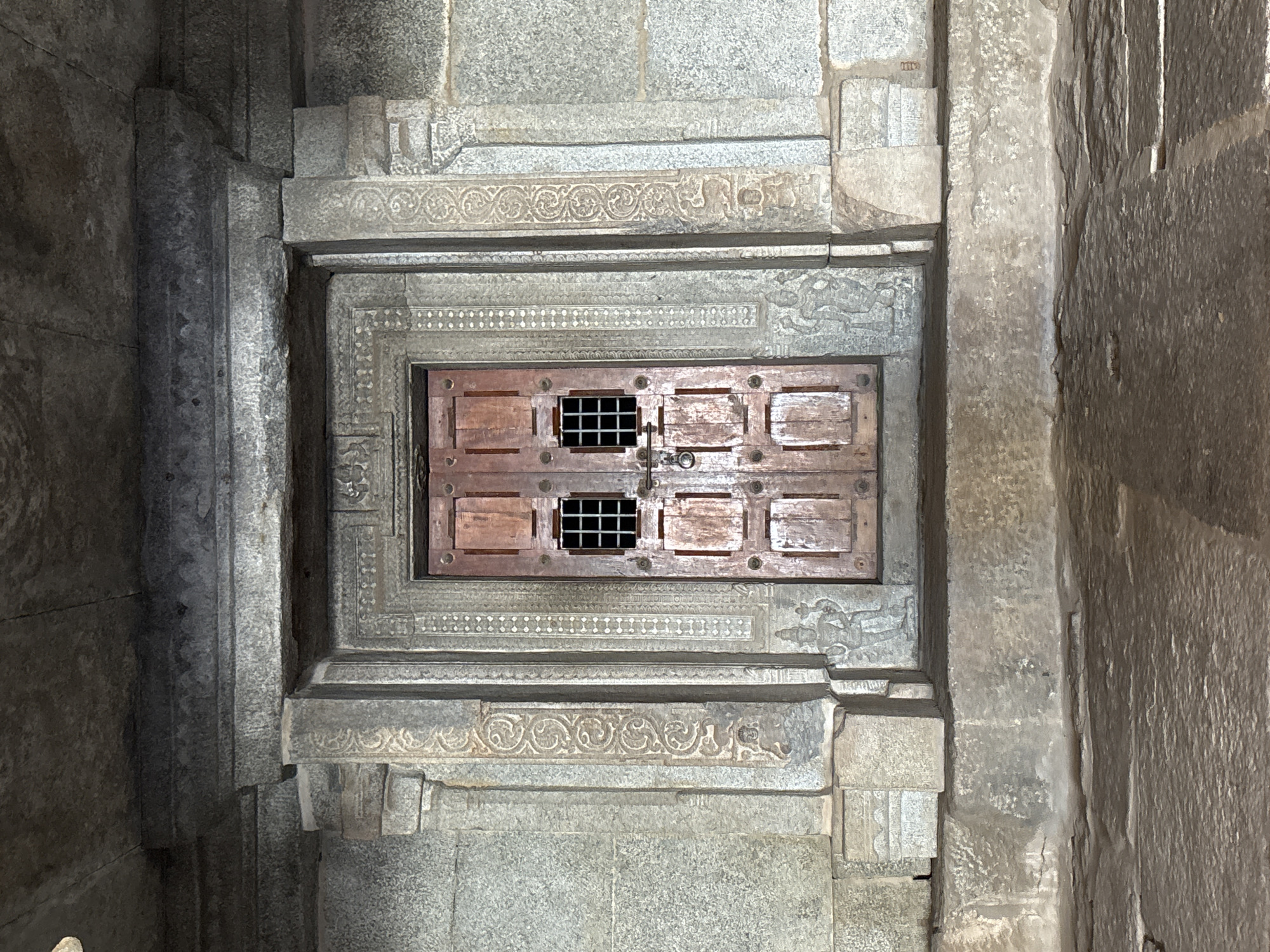 The image shows a detailed view of a wooden door with metal grating set within an ornate stone frame, likely part of a structure in Kamalapura, India. The door is composed of individual panels, secured with a visible latch, and set within a stone recess with multiple levels of carvings and moldings. The surrounding stone walls exhibit a rough texture. No people or other subjects are visible.