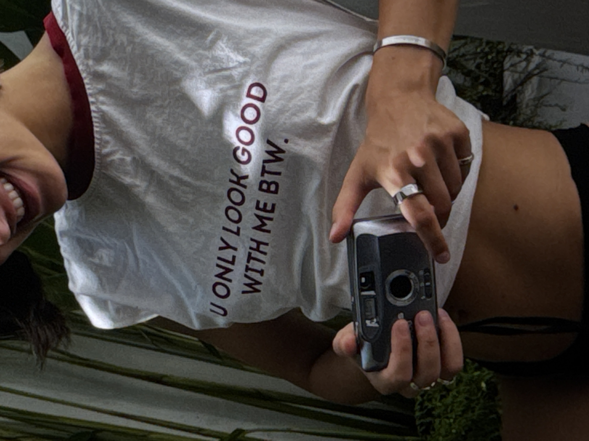 The media file shows a close-up, high-angle, vertical shot of a person's torso and arm. The person is wearing a white t-shirt with maroon text that reads "U ONLY LOOK GOOD WITH ME BTW." They are also wearing a silver bracelet and two silver rings on their fingers. The person is holding a black digital camera with their right hand, and their left arm is bent. Their mouth is open in a smile, revealing teeth. The background appears to be an outdoor setting with green foliage. The lighting suggests it is daytime. The visible text is a playful, possibly ironic, statement.
