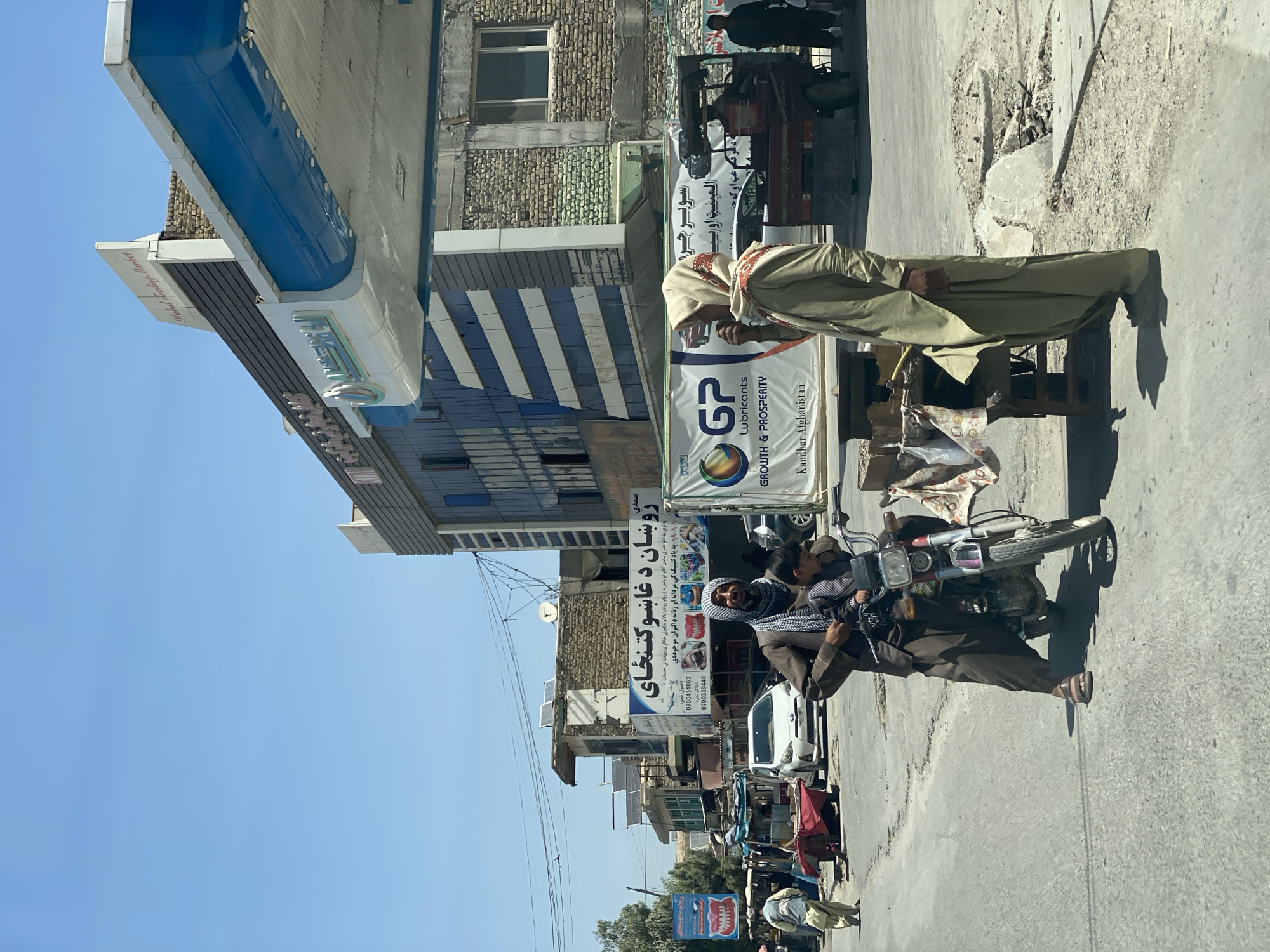 In Kandahar, Afghanistan, a sunny street scene unfolds with several individuals and vehicles. In the foreground, a man wearing traditional clothing and a headscarf stands next to a motorcycle, with a young boy seated behind him. To the right, another man, also in traditional attire and headscarf, stands near a small wooden table with some items on it, possibly engaged in a transaction or conversation. Behind them, a large banner displays the logo of "GP Lubricants" with the text "GROWTH & PROSPERITY" and "Kandhar Afghanistan."
Further back, a white car is parked on the street, and several other vehicles, including a truck and a three-wheeled vehicle, are visible. Buildings with a mix of modern and traditional architecture line the street, some adorned with signs in Arabic script. The environment appears to be a busy commercial area, with a clear blue sky overhead. The road surface shows signs of wear and tear, with some patches of dirt and debris. The overall impression is one of daily life continuing in an urban setting.