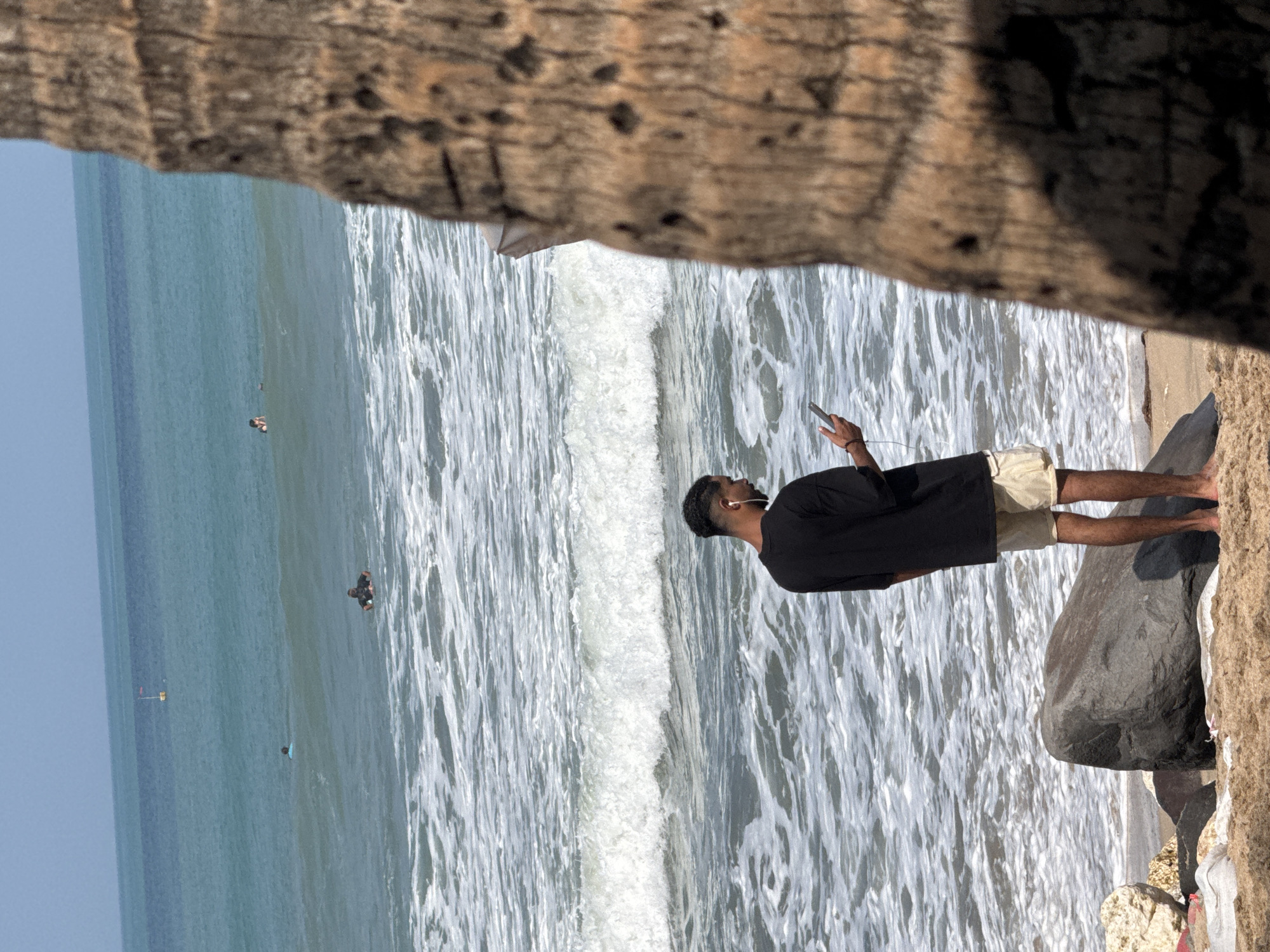 A man stands on a sandy beach in Kuta, Indonesia, facing the ocean. He is wearing a black t-shirt and light-colored shorts, with white earbuds in his ears and holding a smartphone in his right hand. The ocean waves are breaking on the shore, and several people are visible in the water, some appearing to be surfing. A large palm tree trunk is prominently featured in the foreground on the right side of the image, obscuring part of the scene. The sky is clear and blue.