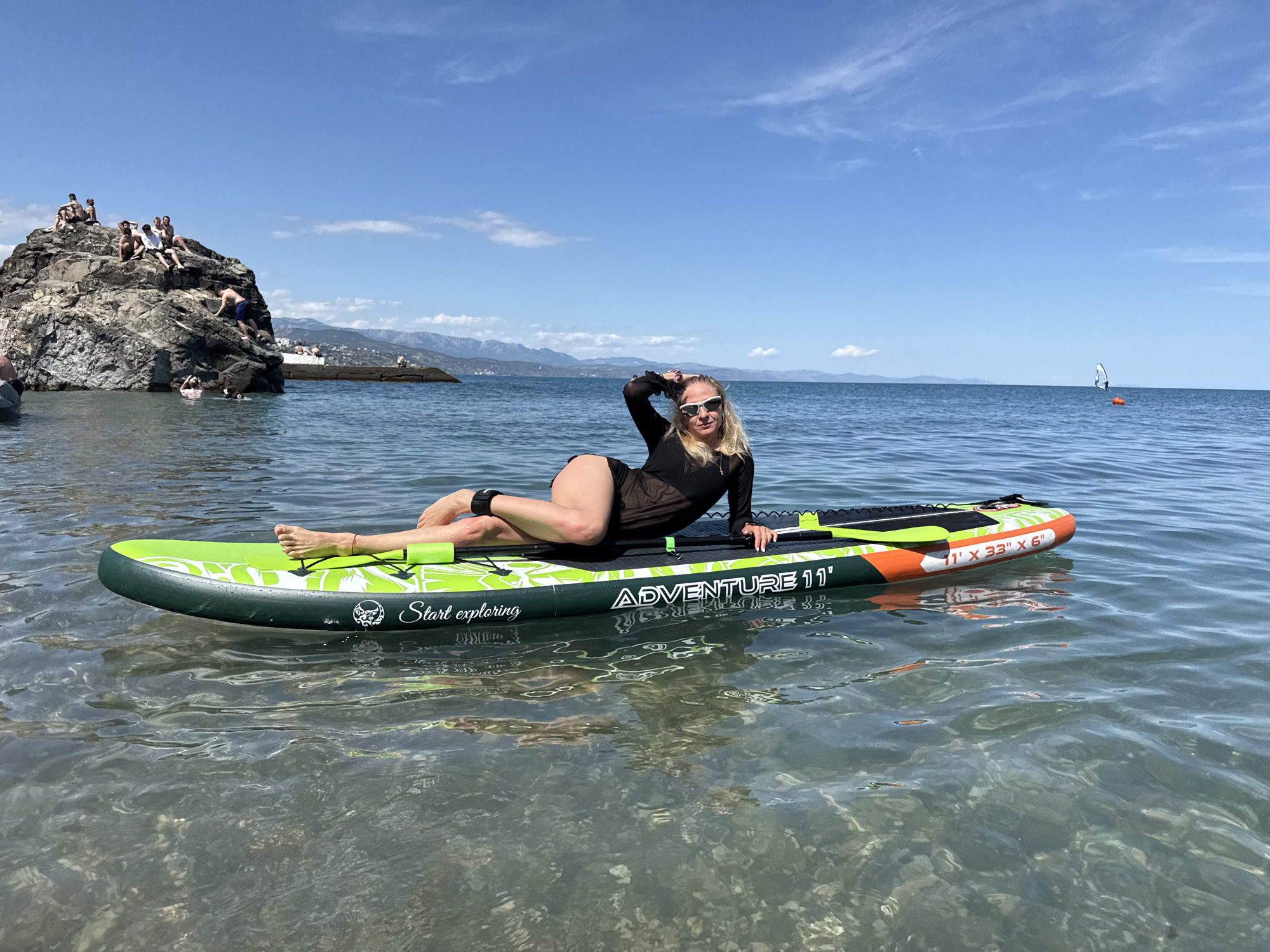 A woman with blonde hair and sunglasses is posing on a green and orange paddleboard in the ocean. She is wearing a black sheer swimsuit. The water is clear and calm, with sunlight reflecting on the surface. In the background, there is a rocky outcropping with several people sitting on top. Beyond that, there are mountains and a clear blue sky with some scattered clouds. The overall scene suggests a warm, sunny day at the beach, likely in the afternoon. The paddleboard has the text "Start exploring" and "ADVENTURE 11'" visible on its side. A windsurfer can be seen in the distance.