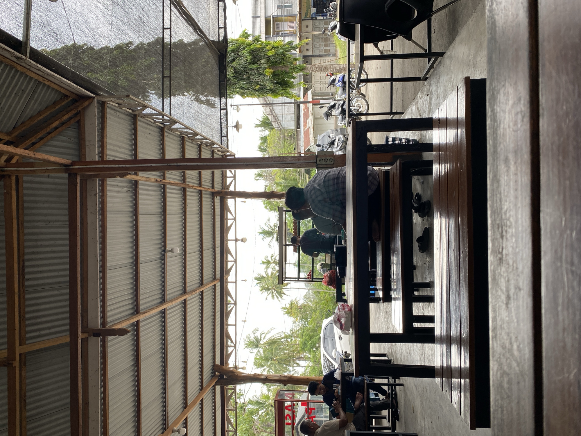 This is a photograph of an outdoor eatery in Cotgadong, Indonesia. The eatery is covered by a corrugated metal roof supported by wooden beams. In the foreground, a wooden table and bench are prominently featured, suggesting a seating area. Beyond this, several other tables and benches are visible, some occupied by people.
Several individuals are seated at the tables, engaged in dining or conversation. On the left side, two men are seated at a table. One man is wearing a light-colored shirt and appears to be looking down at something on the table. The other man, wearing a dark shirt, is looking towards the left. Further back, three men are seated at another table. One man in a plaid shirt is facing away from the camera, while two other men are partially visible. A food stall or counter is also visible in the background, with a person preparing food.
The environment outside the eatery is characterized by palm trees and other greenery, indicating a tropical or subtropical climate. Several motorcycles are parked outside, and a white car is visible in the distance. Buildings with balconies and windows are also present in the background, suggesting a residential or commercial area. The ground is paved with concrete. A mesh-like netting is draped over part of the structure, possibly for shade or protection. The overall scene depicts a casual dining establishment in a somewhat rural or semi-urban setting in Indonesia.