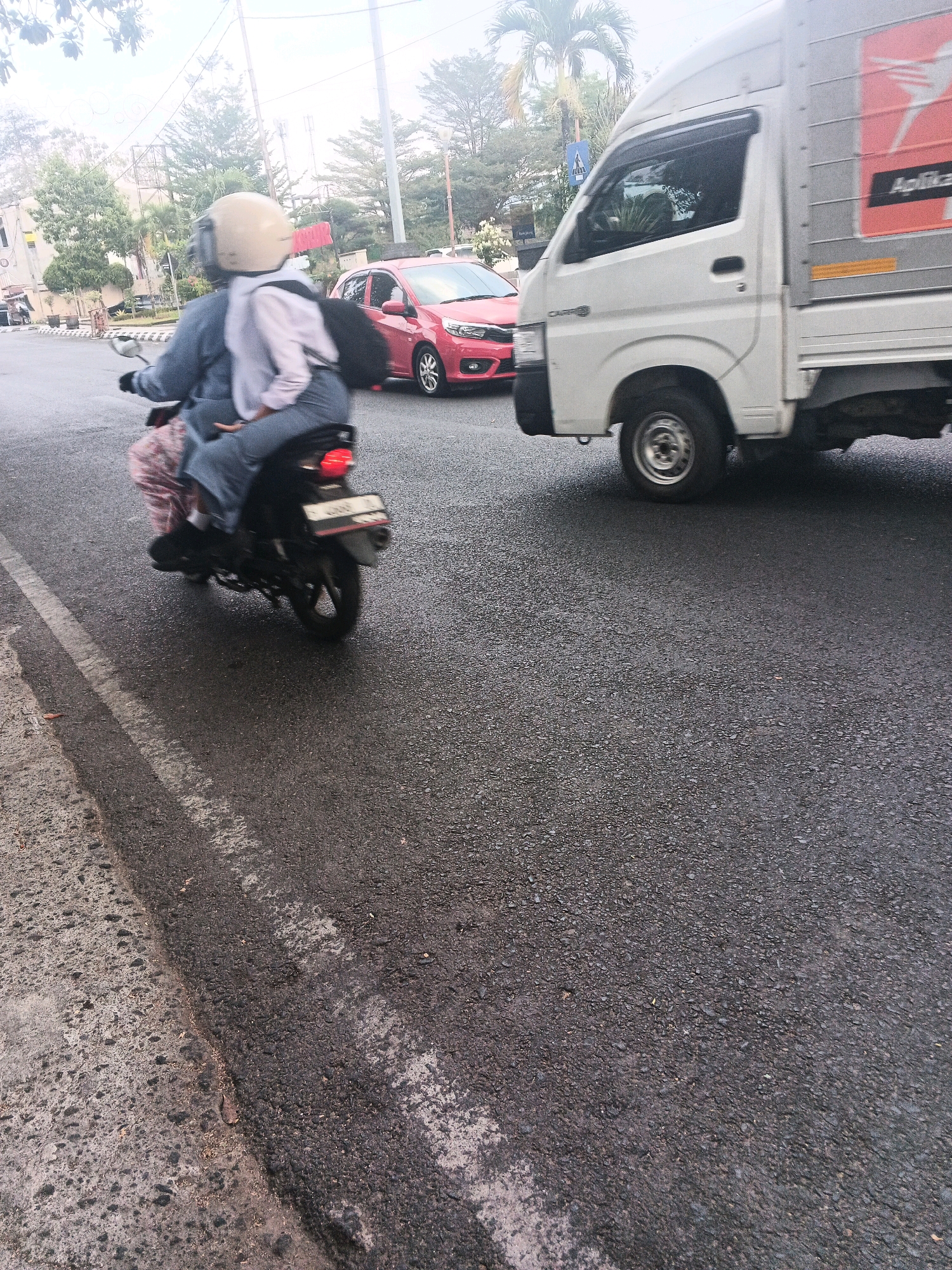 In Sidomulyo, Indonesia, on a wet asphalt road, two individuals are riding a black motorcycle. The person in the front, wearing a light-colored helmet, is operating the vehicle. The person behind them is wearing a light-colored hijab and a blue top, with their arms around the driver. The motorcycle is in motion, and its rear license plate is visible. To the right of the motorcycle, a white truck with a silver cargo box is also in motion. The truck has a red and orange logo on its side. In the background, a red car is parked on the side of the road. Trees and utility poles are visible in the distance, suggesting an urban or semi-urban environment. The road surface shows signs of being recently wet, with reflections visible on the asphalt.