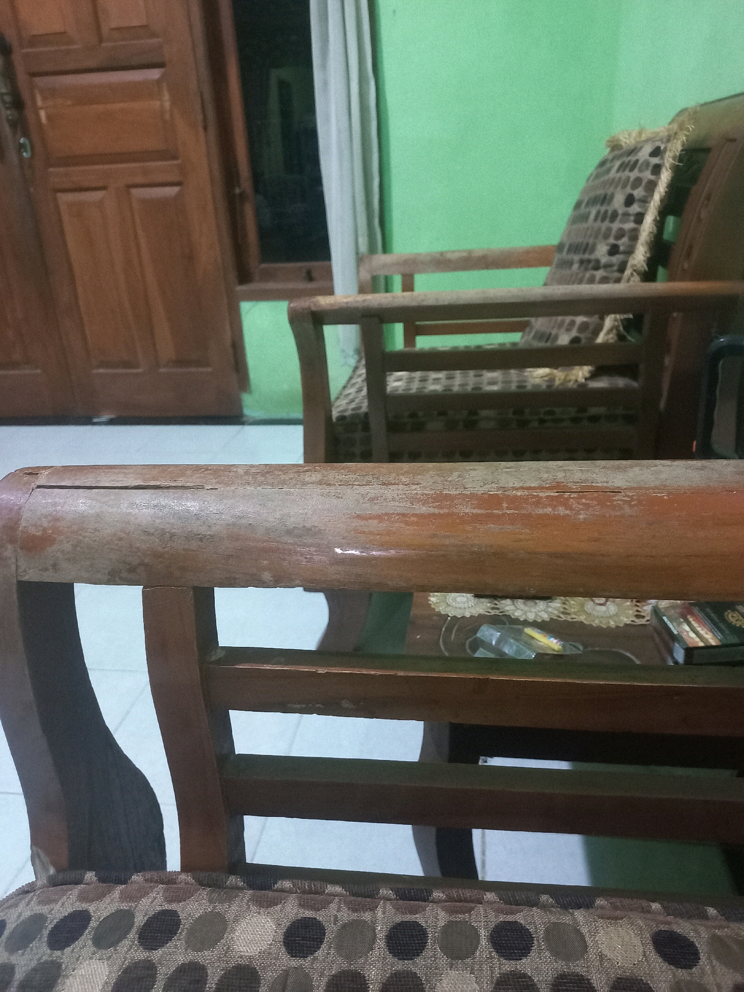 The image depicts the interior of a room, likely a living area, in Sukoharjo, Indonesia. In the foreground, the armrest of a wooden chair with a patterned cushion is visible. The chair appears to be made of dark wood and has a worn finish, suggesting it might be old or well-used. The cushion has a repeating pattern of circles in various shades of brown and gray against a lighter background.
Behind the foreground chair, another similar wooden chair is partially visible. This chair has a pillow with a geometric pattern and frayed edges. The pillow's design consists of a grid of small, dark squares or rectangles on a lighter, textured fabric.
In the background, a large wooden door with paneled sections is prominent on the left side of the frame. The door has a traditional design with distinct raised panels. To the right of the door, a wall painted a light green color is visible. A sheer white curtain hangs near the wall.
The overall impression is that of a simple, domestic setting, possibly a home or a small establishment in Sukoharjo. The lighting is dim, contributing to a somewhat subdued atmosphere. There are no people present in the image, and no specific actions or interactions are being depicted. The focus is on the furniture and the architectural elements of the room.