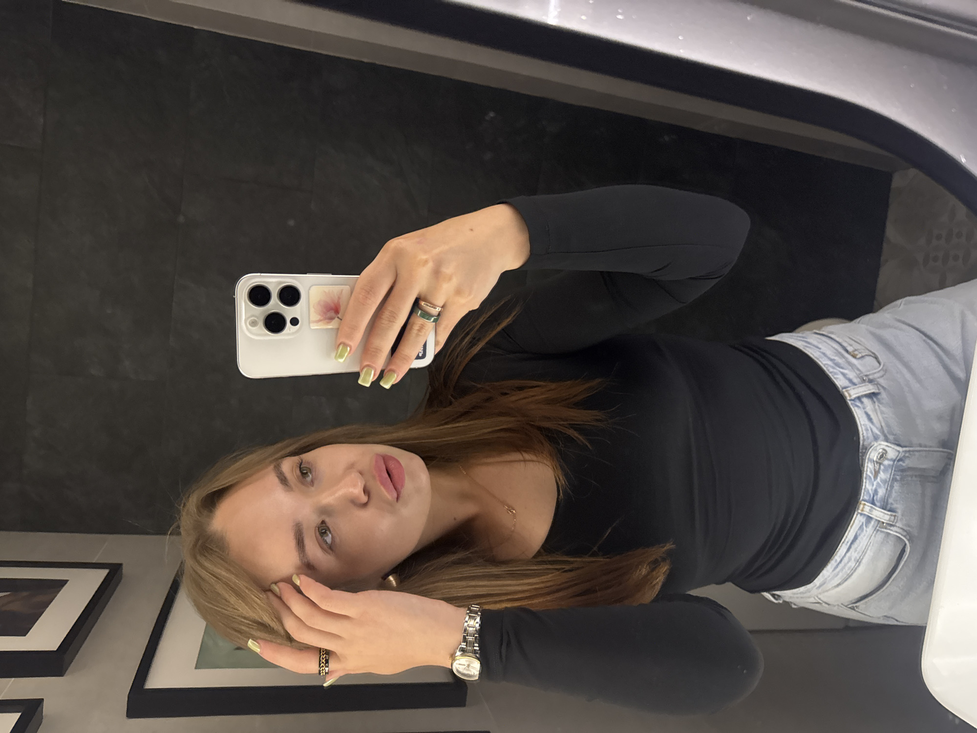 A young Caucasian woman with long, light brown hair is taking a mirror selfie in a dimly lit, modern bathroom. She is wearing a black long-sleeved shirt and light-wash jeans. She has a watch on her left wrist and several rings on her fingers. Her nails are painted a light green color. The background shows dark tiled walls and framed artwork. A white sink is visible in the foreground. The image appears to have been taken in a bathroom in Chaikovsky, Russia.