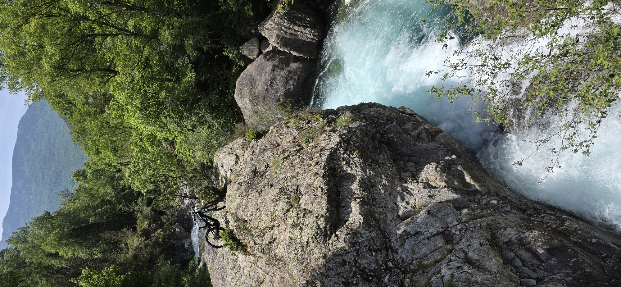 The image shows a mountain bike positioned on a rocky cliff edge, seemingly parked to overlook a rapid river. The cliff face is composed of layered gray and tan rock with patches of green moss. The river is a churning mix of turquoise and white water, suggesting a powerful current. Green foliage, including trees and shrubs, surrounds the rocky area. A mountain range is visible in the background. The location is Sahún, Spain.