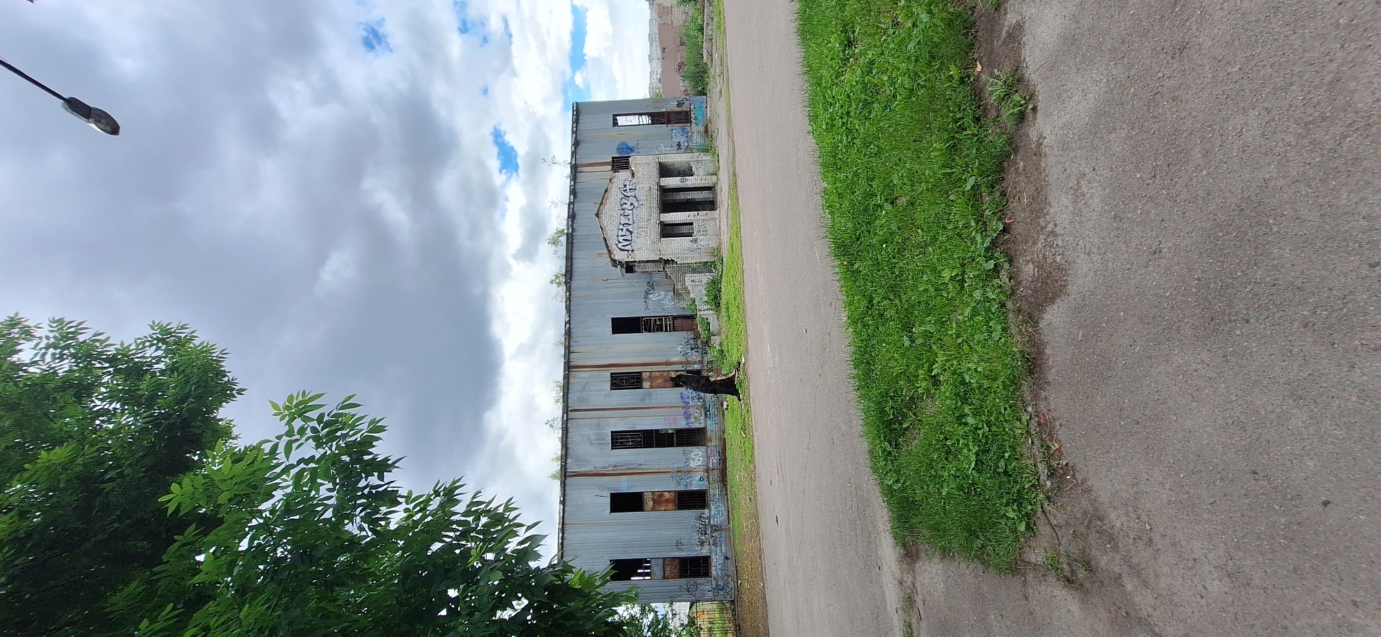 The image depicts an abandoned, graffiti-covered building in Bila Tserkva, Ukraine. The building is made of corrugated metal and brick, with many broken windows. Vegetation is growing on the roof and around the structure, suggesting neglect and the passage of time. A paved path runs alongside the building, bordered by a strip of grass. In the foreground, lush green trees with visible leaves frame the left side of the image. The sky is overcast with scattered clouds, indicating a cloudy day. The overall mood is one of decay and abandonment. There is no visible activity taking place, and no people are present. Text is visible in the form of graffiti on the building's exterior.