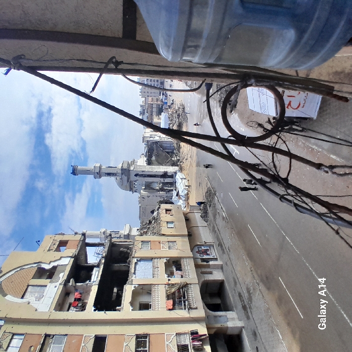 The image presents a severely damaged urban landscape in Khan Yunis, Palestinian Territory, viewed from an elevated perspective, likely a balcony. In the background, a large mosque is heavily damaged, with significant portions of its structure collapsed into rubble, though its central minaret stands largely intact. To the left of the mosque, a multi-story building shows extensive structural damage, with several floors compromised, exposed interiors, and debris hanging from balconies. Patches of laundry are visible on one of the damaged balconies.
An asphalt road, appearing wet in sections and marked with white lane lines, runs through the mid-ground. Piles of rubble and debris line the sides of the road near the damaged structures. Approximately four individuals are standing on the road in the mid-left, while other people are visible walking further down the street towards the background and near the mosque's remains. To the right of the mosque, amidst further rubble, several white tent-like structures or makeshift shelters are present.
In the immediate foreground on the right, a large, translucent blue water cooler bottle is partially visible. Dark, possibly rusted, metal balcony railings and associated wiring diagonally traverse the right side of the frame, indicating the vantage point. The sky overhead is partly cloudy with visible patches of blue.