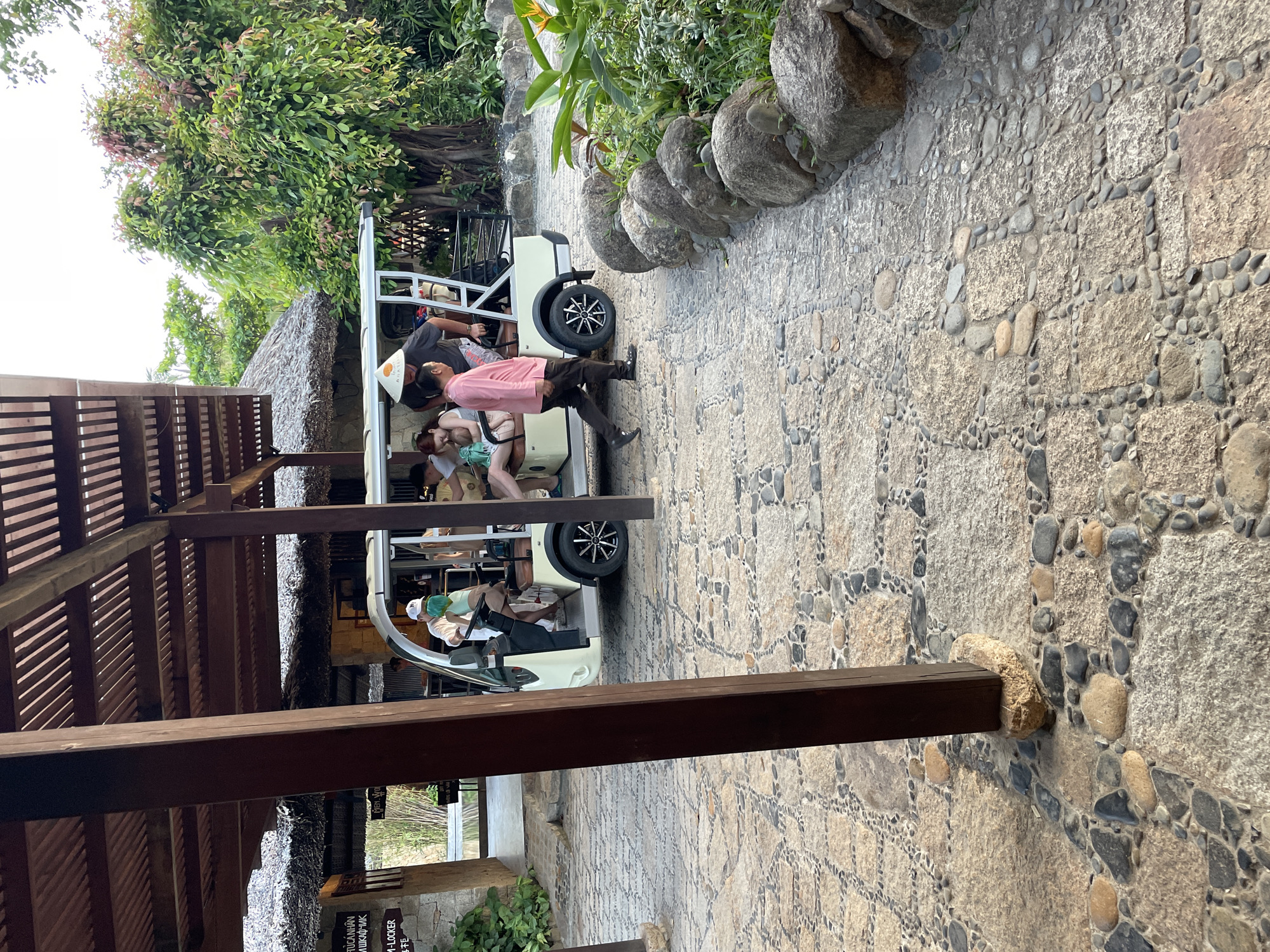 The image shows a golf cart filled with people traveling on a stone-paved pathway in what appears to be a resort or park setting. The location is Nha Trang, Vietnam. The golf cart is white and has a simple frame structure. Several people are inside, including a driver wearing a face mask. One man, wearing a pink shirt and dark pants, is partially outside the cart, seemingly stepping out or assisting with the vehicle's movement. The pathway is bordered by a row of large stones on one side. A thatched-roof structure with wooden support beams is visible on the left. Lush greenery and trees surround the pathway, contributing to a tropical environment.