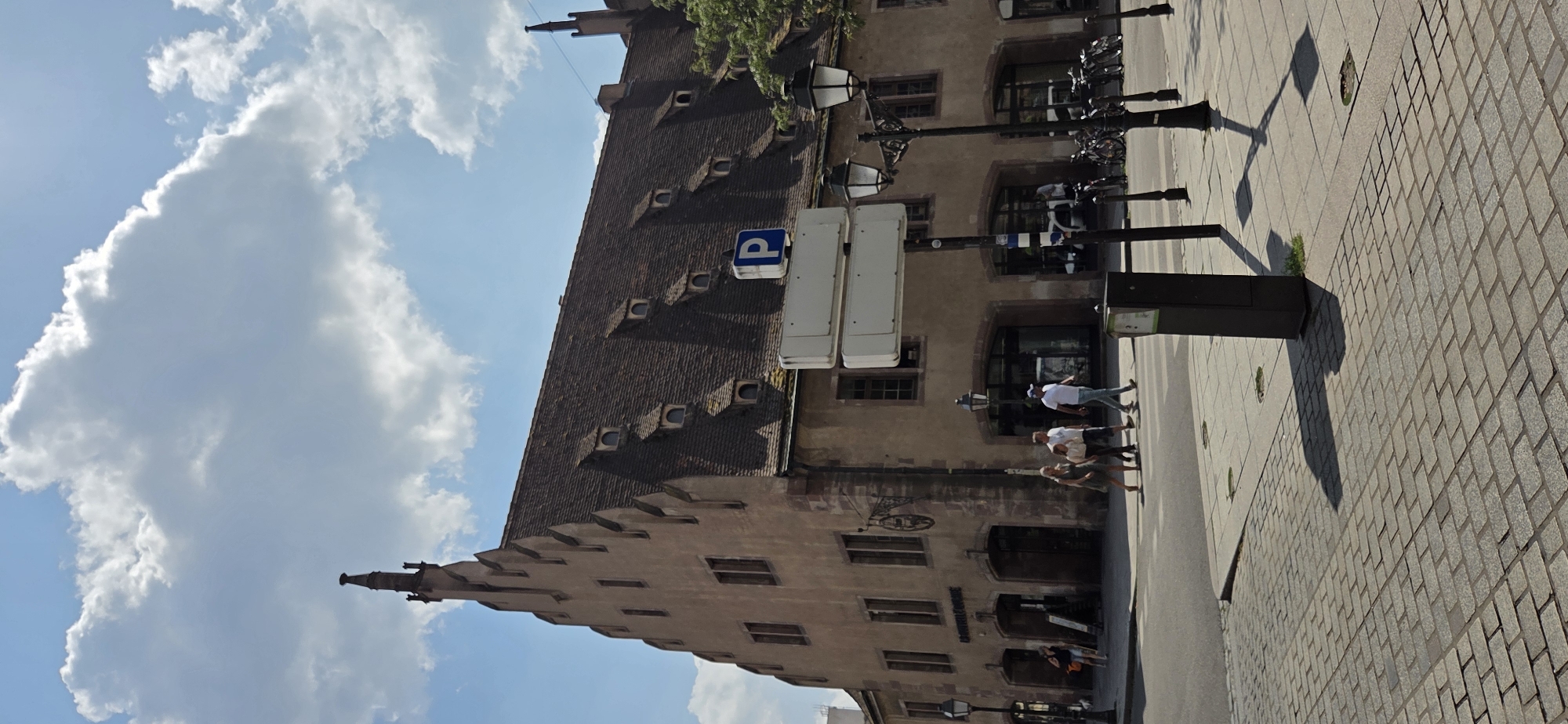The image depicts a street scene in Strasbourg, France, under a bright, partly cloudy sky. A tall, historic building with a steep, tiled roof and multiple dormer windows dominates the right side of the frame. The building's facade is made of a light-colored material, possibly stone or stucco, and features several arched entrances and windows. A sign with a blue "P" indicates parking. In front of the building, a group of people are crossing the street at a marked crosswalk. One person is walking a dog. Streetlights and parked bicycles are also visible. The ground is paved with cobblestones and square tiles. The overall impression is that of a picturesque, European urban environment on a sunny day.