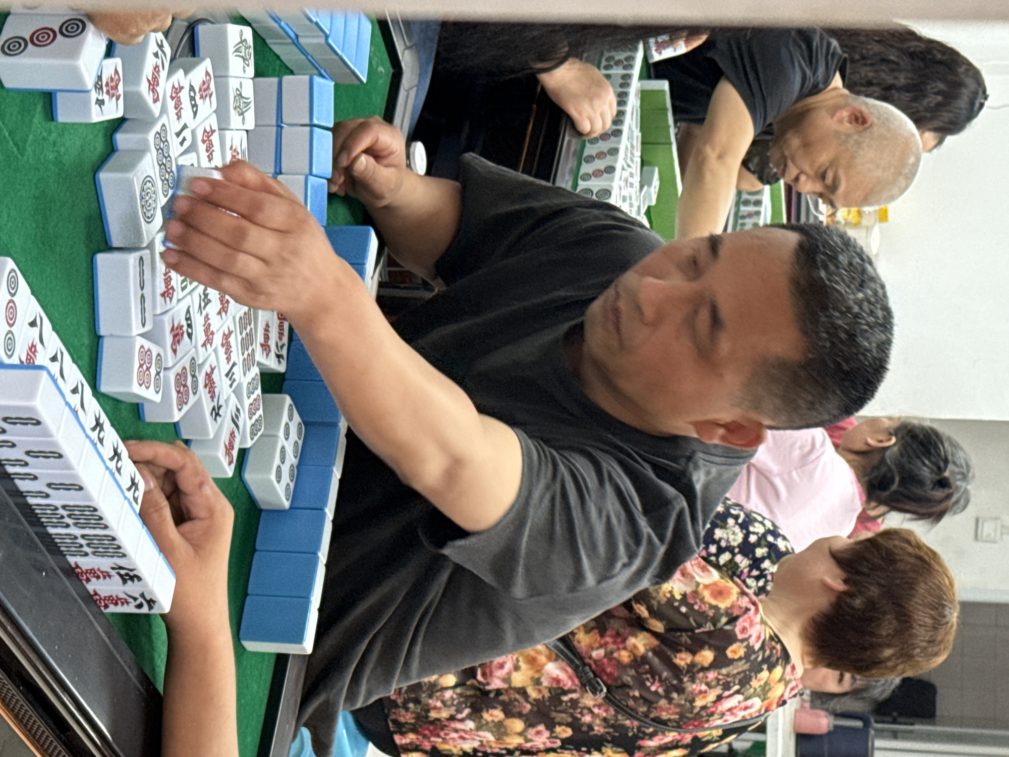 A group of people are playing Mahjong, a tile-based game, at a table covered with a green cloth. The people appear to be middle-aged or older and are gathered around the table. The foreground is filled with mahjong tiles in various arrangements. Based on the location provided (Chongqing High-tech Zone, China), this is likely a social gathering centered around a popular game in Chinese culture.