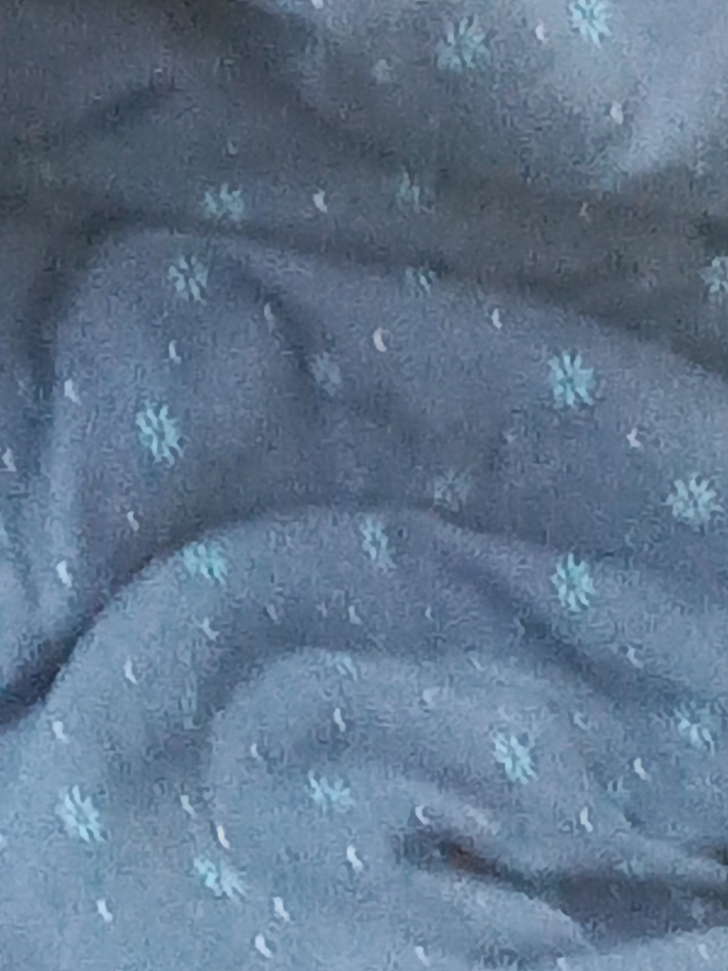 The image shows a close-up of a blue fabric with a pattern of small, stylized blue flowers and white curved dashes. The fabric is wrinkled and textured. No people, subjects, actions, or interactions are visible. The environment is limited to the fabric itself. No city or location is discernible.