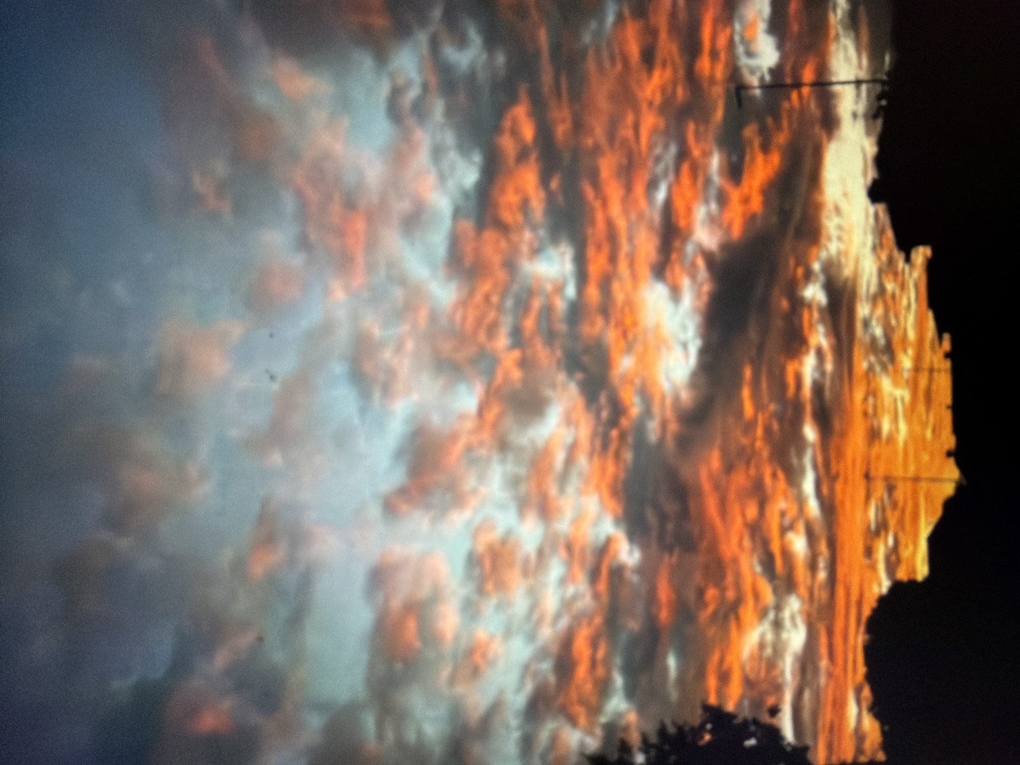 This media file is a photograph of a dramatic sky, likely taken at sunset or sunrise due to the vibrant orange and red hues of the clouds. The clouds are the primary subject, appearing turbulent and illuminated by a low-angled sun. Some clouds are a bright, fiery orange, while others are softer shades of pink and white. There are also darker, more ominous clouds present, creating a stark contrast.
On the right side of the image, a dark silhouette of a person or a structure is visible against the brightly lit sky. This silhouette is jagged and appears to be against a dark background, suggesting it might be a rooftop or the edge of a building. A thin, dark horizontal line can be seen extending across the sky from the silhouette, which could be a wire or part of a structure.
The overall impression is one of intense natural beauty, with a sense of drama and perhaps a touch of foreboding due to the dark silhouettes and the intensity of the colors. The image is somewhat grainy, suggesting it might have been taken in low light conditions or with an older camera. There is no visible text in the image.