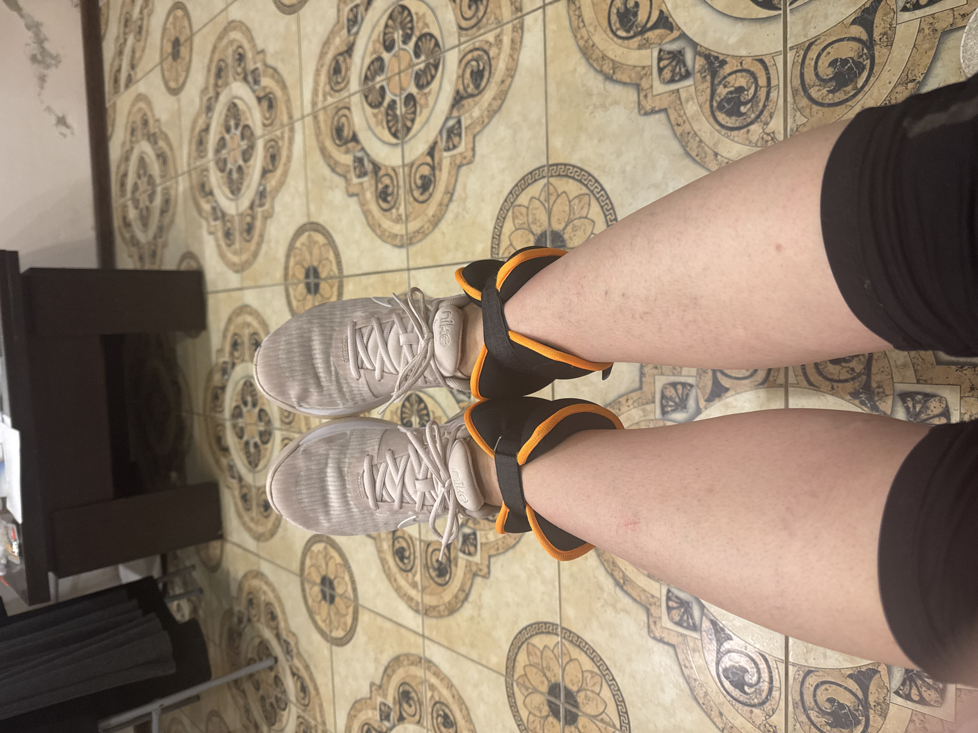 The image shows a person's legs and feet wearing gray athletic shoes and black and orange ankle weights. The person is standing on a tiled floor with a patterned design. In the background, there is a dark-colored piece of furniture or equipment. The specific location in Sochi, Russia, cannot be determined from the image.