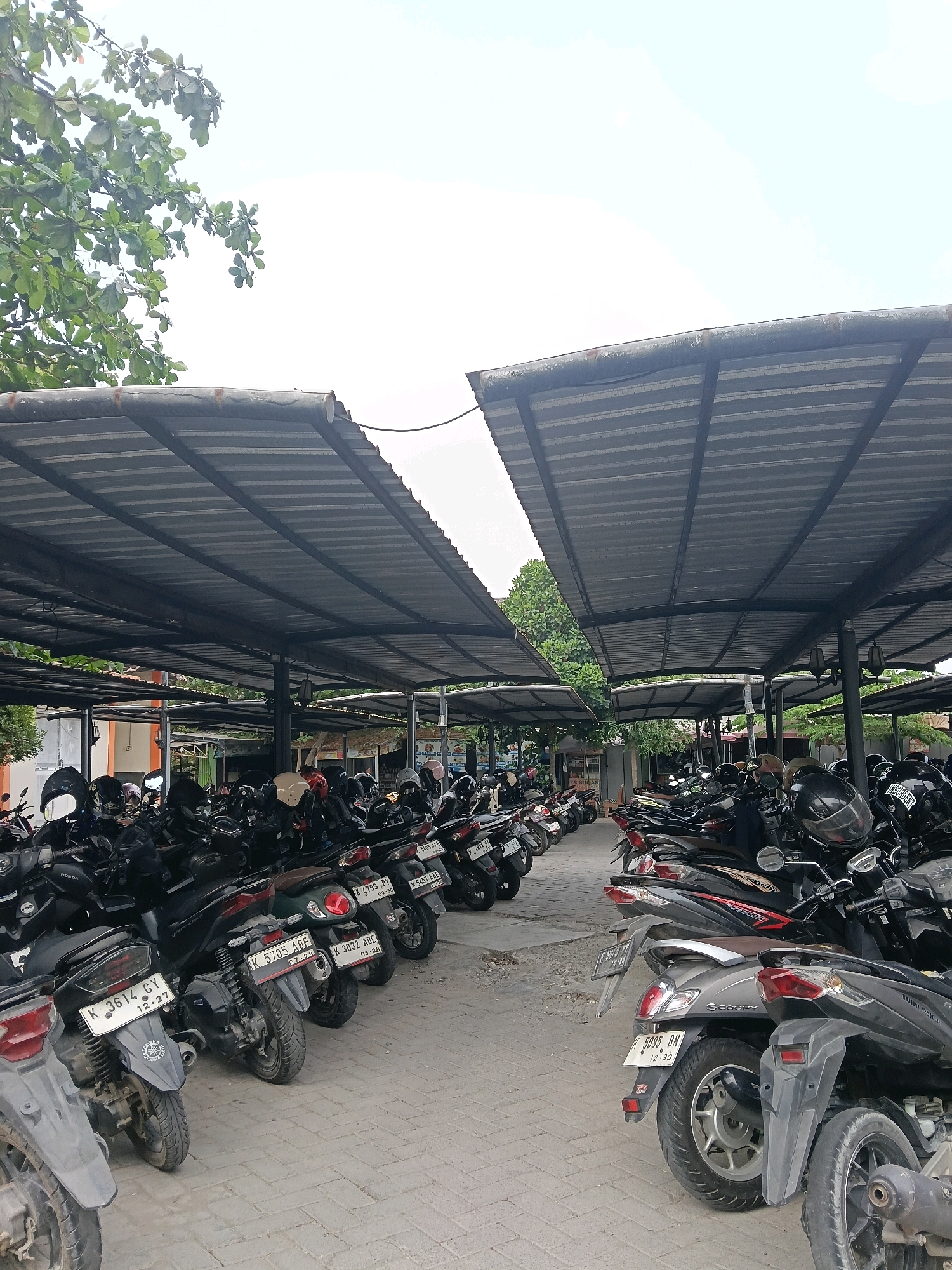 The image captures a covered outdoor parking lot filled with numerous motorcycles in Balun, Indonesia. The lot is covered by two large, weathered metal roofs supported by dark metal pillars. Rows of parked scooters and motorcycles, primarily dark-colored, fill the space. Many of the motorcycles have helmets on their seats or handlebars. The ground is paved with interlocking gray bricks. Green trees and foliage are visible behind the parking structures, suggesting an outdoor setting. The sky is overcast and bright. The scene appears to be a typical parking area, likely for a business or public facility.