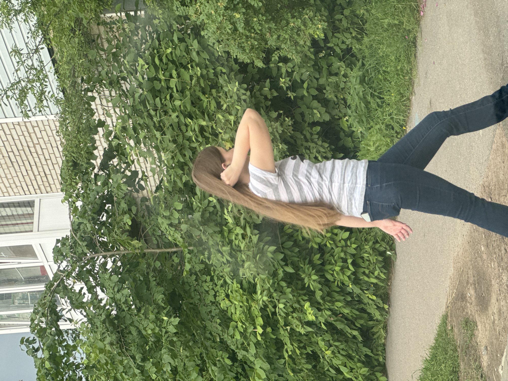The media file shows a person from the side, standing on a paved path next to a dense wall of green foliage. The person has long, light brown hair that cascades down their back, and is wearing a striped t-shirt and dark jeans. Their right arm is bent, with their hand resting near their head, obscuring part of their face. Their left arm is extended downwards, with their hand open. The scene appears to be outdoors, with lush green bushes and grass on one side of the path, and a building with light-colored brick and large windows on the other side. The lighting suggests it is daytime. The location context is Kuzmolovskiy, Russia. There is no visible text in the image.