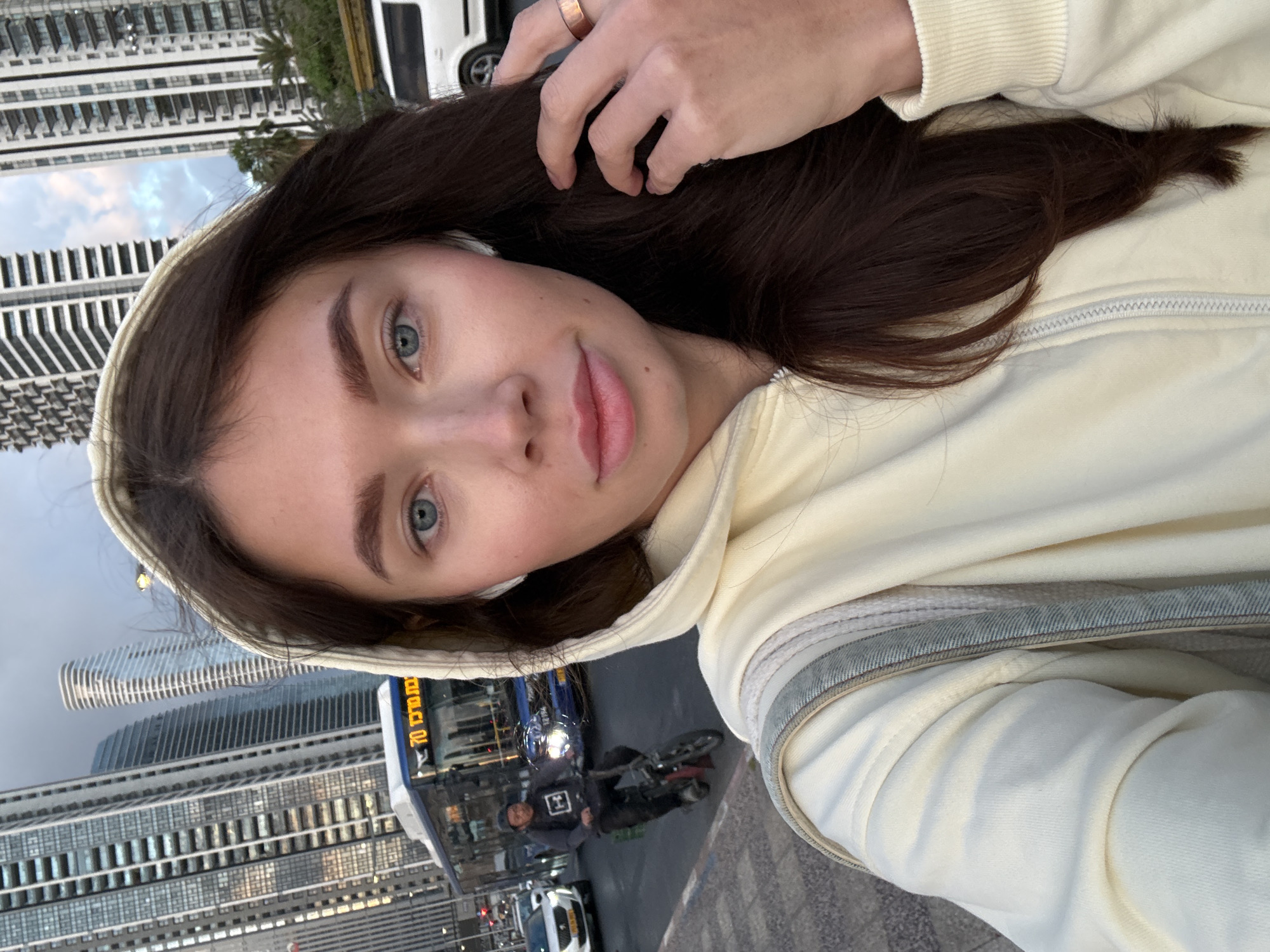 The image, oriented on its side, presents a close-up selfie of a young woman with long, dark hair, striking blue eyes, and a calm, neutral expression. She wears a light, cream-colored hooded sweatshirt, and her right hand, adorned with a ring, is visible near her head.
The background reveals a bustling urban street scene, characteristic of Tel-Aviv, Israel. Tall, modern high-rise buildings with numerous windows line the street, appearing horizontally in the current orientation but vertically in an upright view. The sky above is overcast. On the street below, a blue public transport bus is prominent, displaying the route number "70" and the Hebrew text "מטרופולין" (Metropoline), identifying it as an Israeli bus company. Beside the bus, a person on a motorcycle or scooter is visible, wearing a dark hoodie with a white square logo on the back. Other vehicles, including a white van and another car, are also part of the traffic. A palm tree adds a touch of greenery amidst the urban structures. The subdued lighting suggests an overcast daytime.