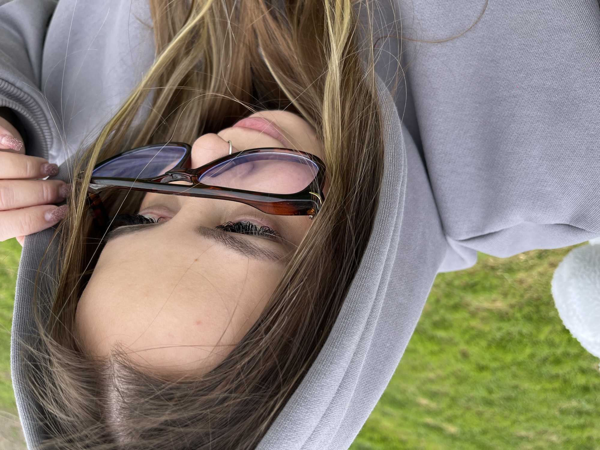 The image is a close-up, high-angle portrait of a person, presented with a clockwise rotation of approximately 90 degrees. The main subject, likely a young female, is wearing a light gray hooded sweatshirt, which covers much of their upper body. Long, multi-toned brown and blonde hair cascades around their face and over the hoodie.
The right side of the subject's face is prominent, including the forehead, a portion of the right eye with striking dark eyelashes, the nose, upper lip, and chin. A small silver hoop nose ring is visible on the right nostril. A pair of dark brown tortoiseshell-patterned glasses rest on the bridge of their nose, with the lenses partially covering the eye and exhibiting a blue anti-reflective tint. The person's left hand is visible on the left side of the frame, with fingers gently holding strands of hair; the nails are painted with a glittery pink polish.
The background is a soft, blurred expanse of green grass, indicating an outdoor environment, possibly a park or field in Port-Katon, Russia. The lighting suggests it is daytime. On the far right edge, a small, out-of-focus white fluffy object is partially visible.
The scene conveys a casual and relaxed mood, depicting the person in an unconventional pose, possibly looking upwards or taking a selfie from an unusual angle. There is no visible text in the image.