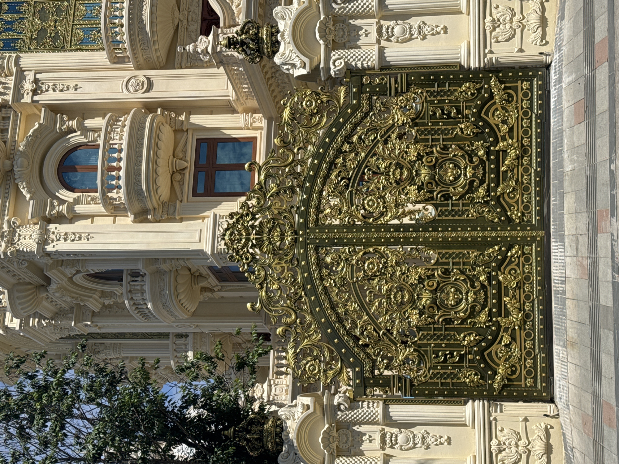 The image shows a large, ornate, dark green gate with intricate gold detailing, likely made of metal. The gate is adorned with elaborate scrollwork, floral motifs, and small, raised studs. It is flanked by cream-colored stone pillars and walls with similar classical architectural ornamentation, including columns, arches, and decorative reliefs. Behind the gate, a section of a building is visible, characterized by its light-colored stone facade, multiple balconies with ornate balustrades, arched windows, and decorative molding. A tree with green leaves is partially visible on the left side of the image. The ground in front of the gate is paved with rectangular tiles in shades of gray and reddish-brown. The scene appears to be a grand entrance, likely to a mansion or a high-end establishment. Based on the architectural style and decorative elements, this is likely the entrance to an opulent mansion located in the city of Da Lat, Vietnam, a city known for its French colonial architecture and grand villas. There are no people visible in the image.