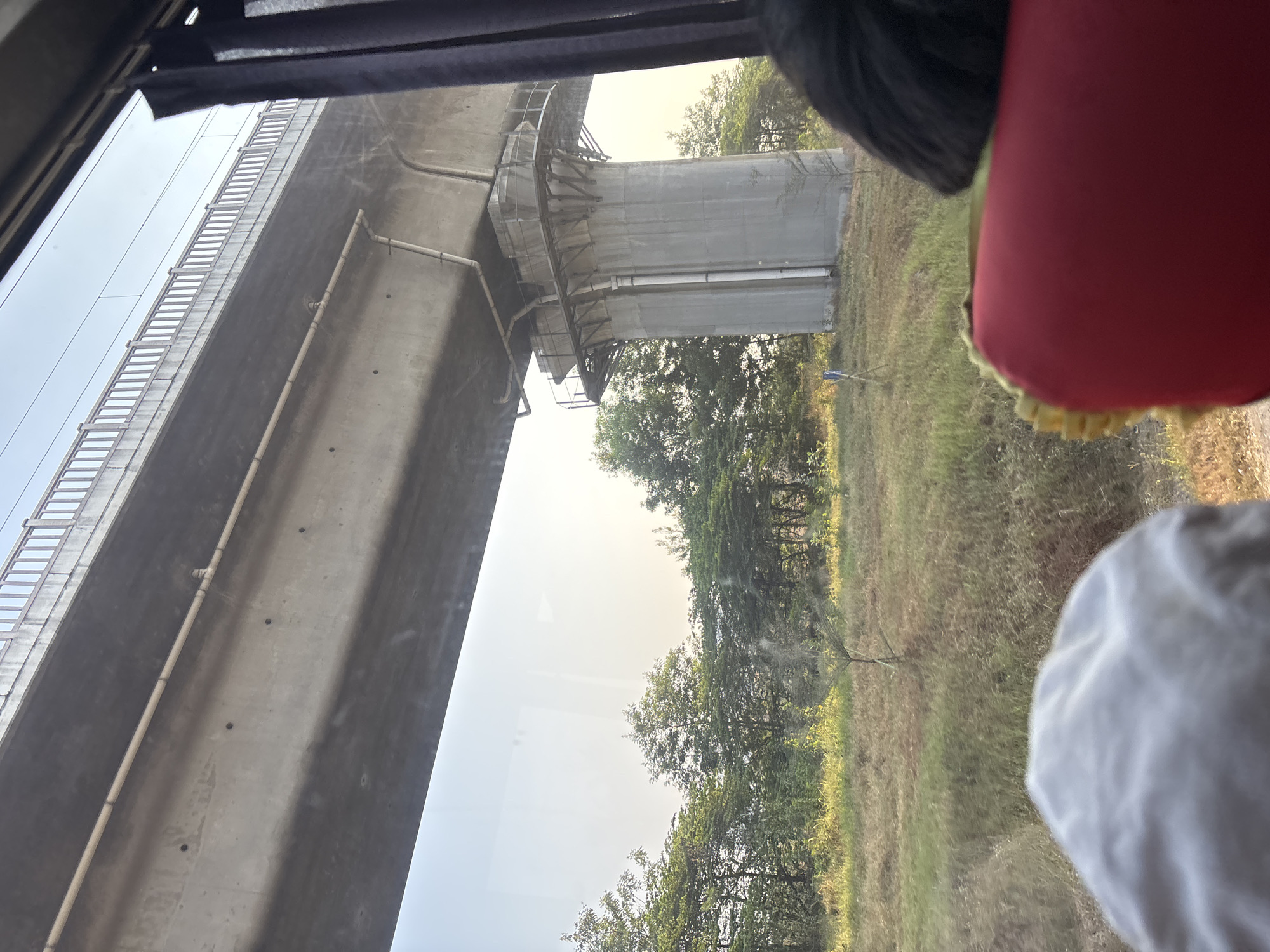 The image shows a view from inside a vehicle looking out at an elevated concrete structure, likely an overpass or bridge, with trees and grassy fields in the background. The overpass has a railing along its top and appears to have some piping attached to its underside. A large concrete pillar supports the overpass. In the foreground, parts of the vehicle's interior are visible, including a red seat with yellow trim and a white cushion. Two individuals are partially visible in the foreground, one with dark hair and another with a white head covering. The scene appears to be in a rural or semi-rural area, possibly on the outskirts of Jayamukti, Indonesia, given the vegetation and the construction of the overpass, which is typical of infrastructure development in the region. The lighting suggests it is daytime, possibly late afternoon, with the sun casting a warm glow on the landscape.
