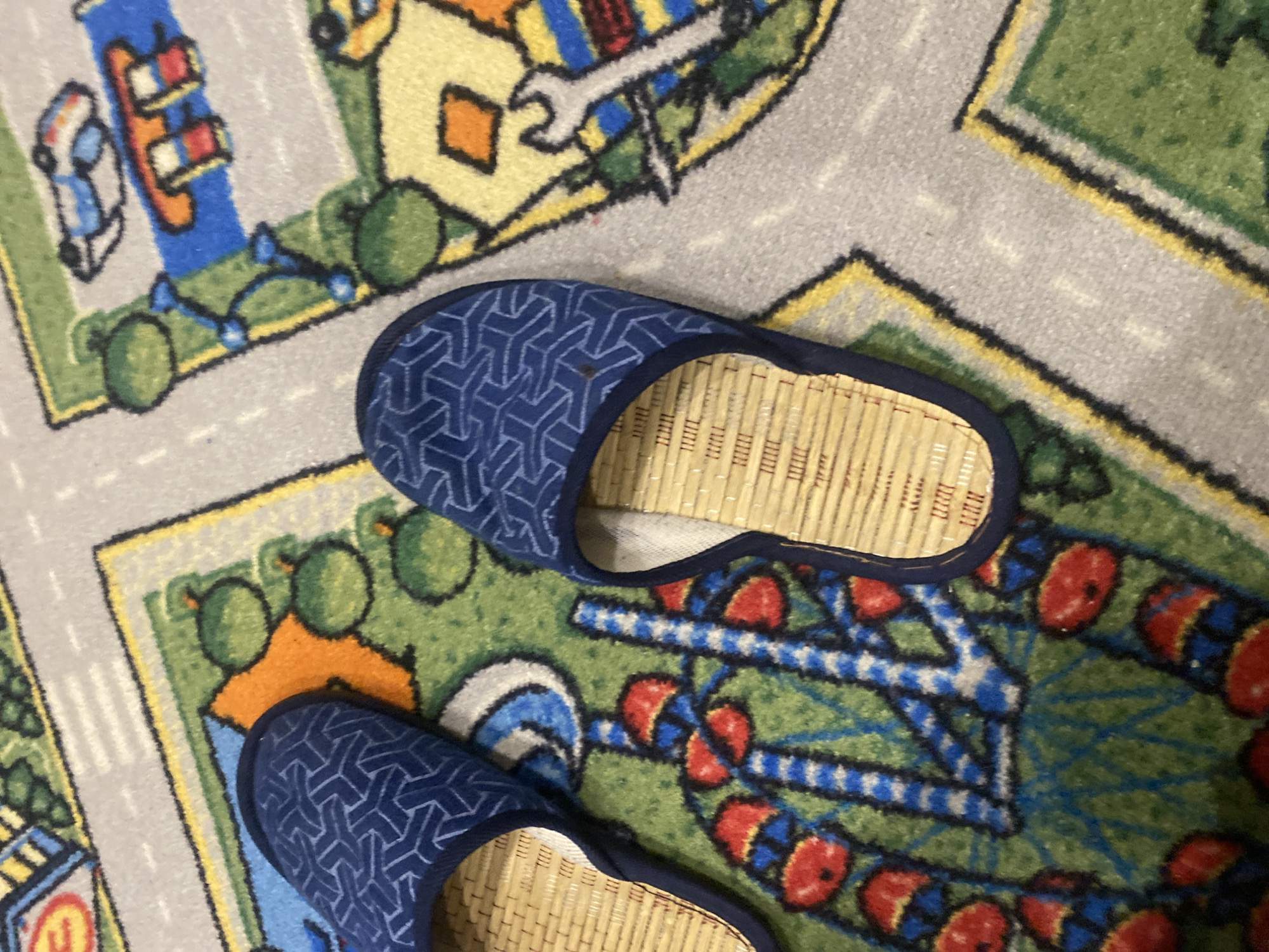 The image shows two blue slippers with a geometric pattern placed on a colorful rug that depicts a cityscape with roads, buildings, and various objects. The slippers have woven natural material insoles. The rug has a textured surface and appears to be a playmat for children, with depictions of toy cars, traffic lights, and other urban elements. The lighting suggests an indoor setting. There is no identifiable information in the image that would allow for its location to be determined to Saratov, Russia.