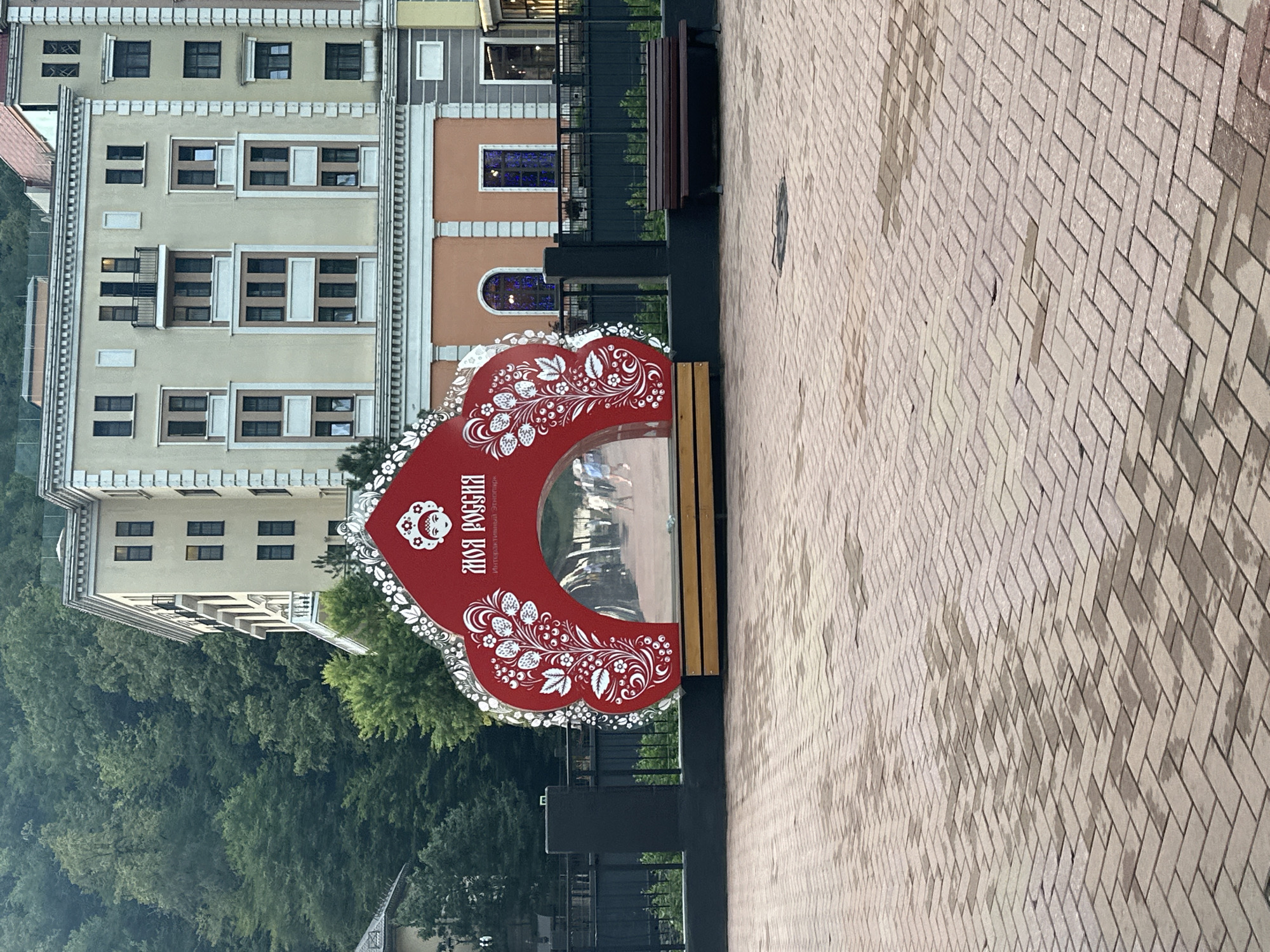 This is a photograph taken outdoors in the city of Estosadok, Russia, likely in a public square or pedestrian area. The foreground consists of a wet, herringbone-patterned brick paving. In the midground, a large, ornate, red, kokoshnik-shaped structure stands, decorated with white floral patterns and a depiction of a woman's face in traditional Russian headwear. The text "МОЯ РОССИЯ" (My Russia) is visible on the structure, along with smaller text underneath. This structure appears to be a decorative installation or photo booth with a mirrored interior, reflecting a pathway and several people walking. To the right of this structure, a dark wooden bench is positioned on a slightly raised platform. Behind the kokoshnik installation, a black metal fence encloses a pathway. Further in the background, several multi-story buildings with pale yellow facades and dark windows are visible, situated against a backdrop of dense green trees on a hillside. The sky is not visible in the frame.