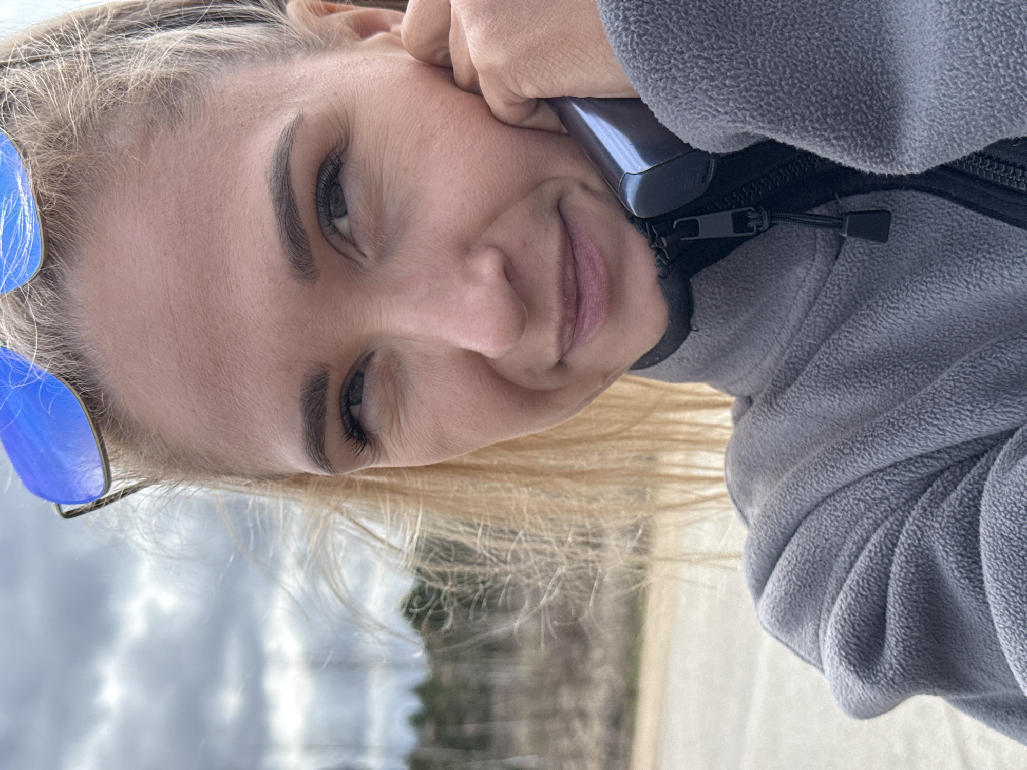 A person with light skin and blonde hair is holding a phone to their ear and smiling. They are wearing a gray fleece jacket with a black zipper. Blue sunglasses are visible on their head. The background shows a street and trees under a cloudy sky. The city of Гремяча, Russia, is not discernible from the image.