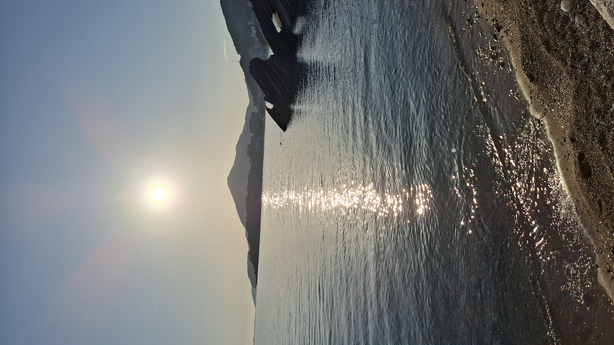 A landscape photograph shows a view from the shore in Boreti, Montenegro, under a bright sun. The scene depicts a clear sky with the sun high above, casting a golden hue on the landscape. A mountainous silhouette stretches along the left side, meeting the calm sea. The water reflects sunlight, creating a glittering effect on the surface. The beach, composed of dark sand and pebbles, borders the sea on the right, marking the transition from water to land. The overall composition suggests a serene and natural setting.