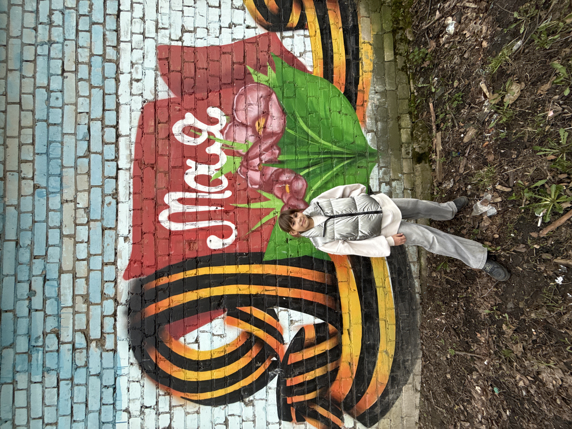 A young person wearing a silver puffer vest and gray pants is lying on a brick mural in Rusanovo, Russia. The mural, painted on a brick wall, is predominantly in shades of red, orange, and black, with accents of green and white. The left side of the wall is painted blue and white, contrasting with the vibrant mural. The right edge of the brick surface is adjacent to an area of dark soil and vegetation. The person is positioned centrally in the frame, with their body mostly on the mural.