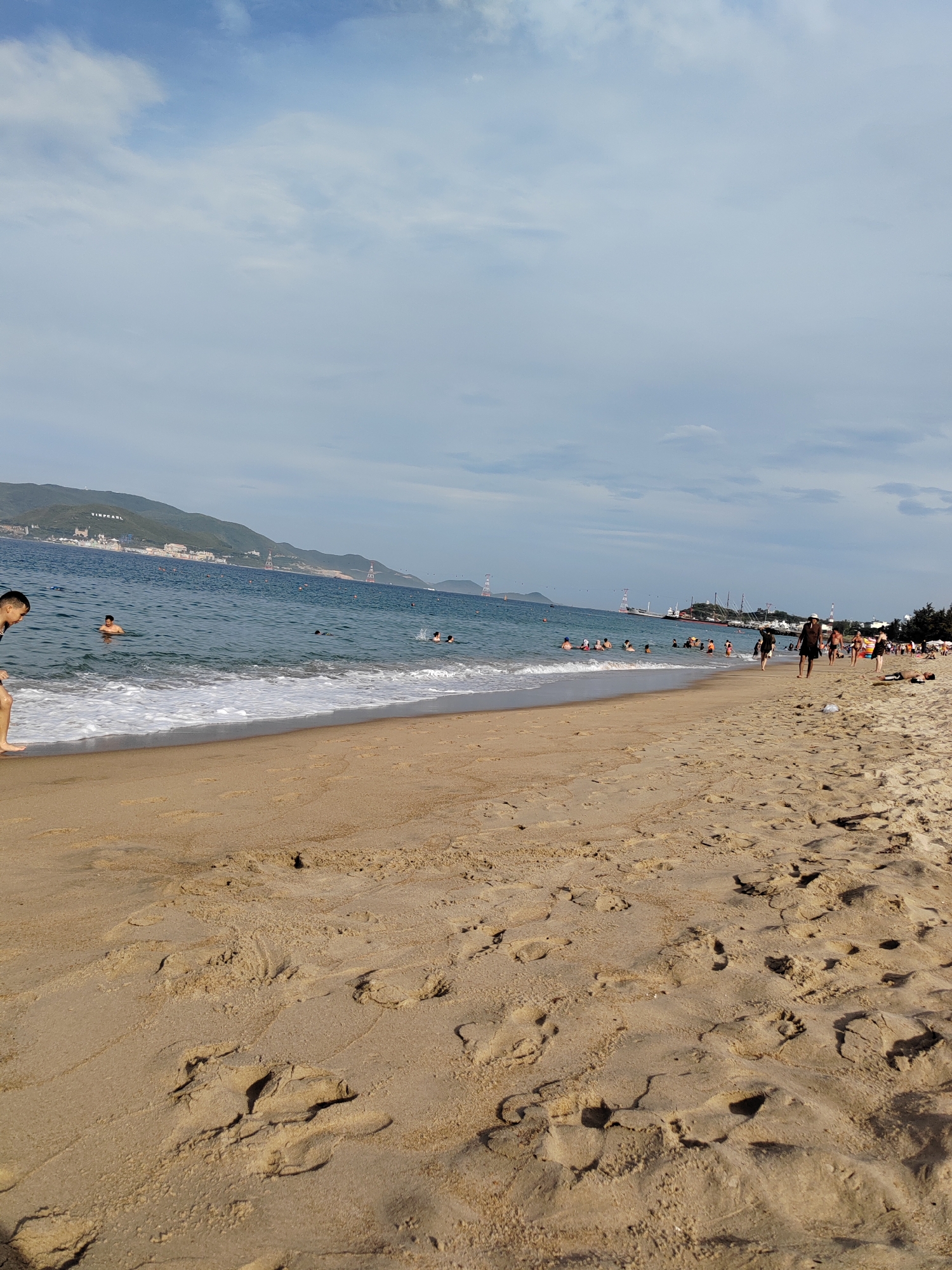 The image depicts a crowded beach in Nha Trang, Vietnam, with numerous people swimming and walking along the shore. The sandy beach is wet near the waterline, indicating recent waves. In the distance, lush green hills rise behind a strip of buildings, suggesting an urban coastal setting. Several boats and a large cable car system are visible on the water and in the sky, respectively, further confirming the location. The sky is partly cloudy, with a mix of blue and white. The overall scene conveys a lively atmosphere on a sunny day at the beach.