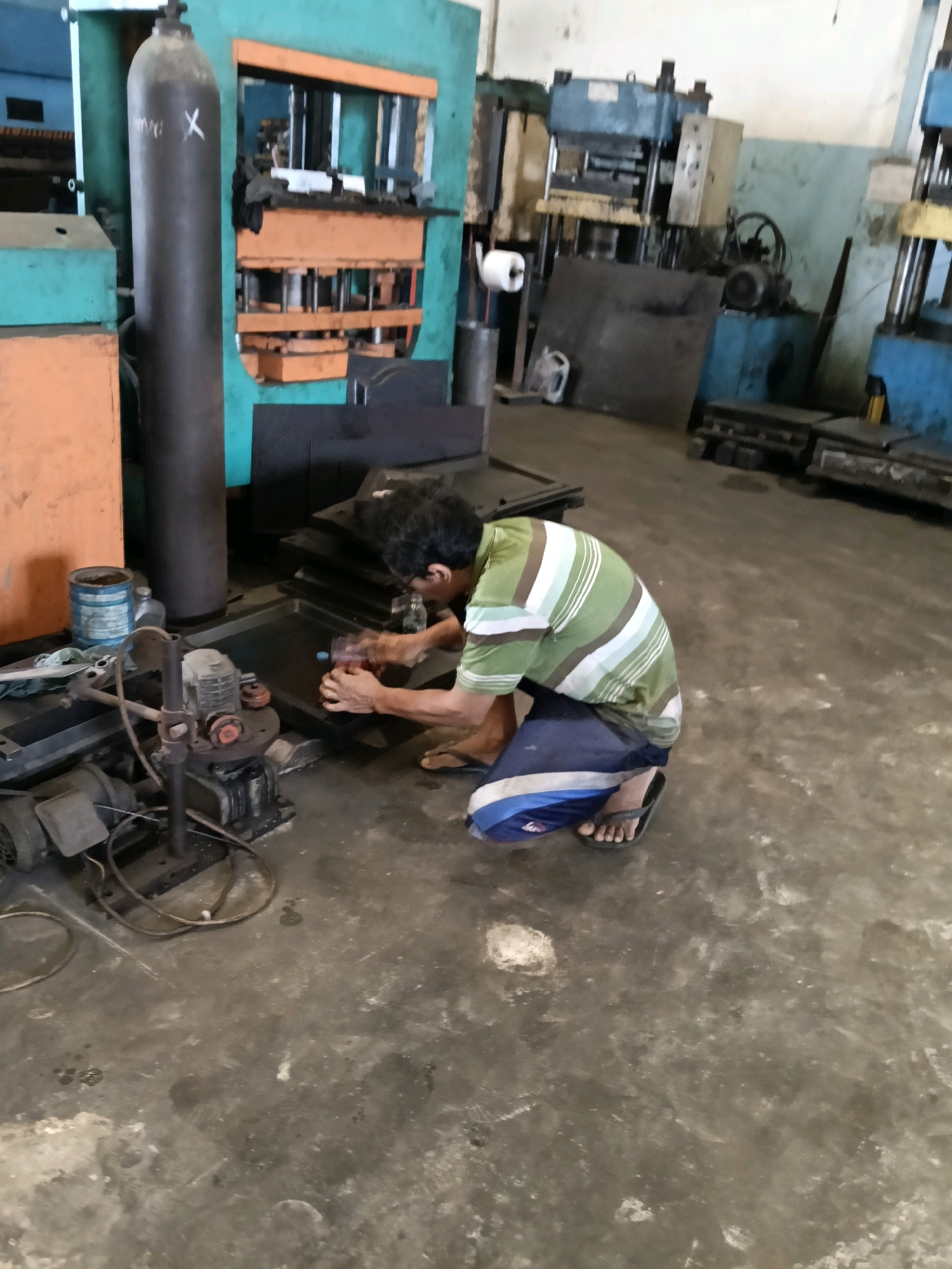 In the city of Belimbing, Indonesia, a man is seen kneeling on the concrete floor of what appears to be a workshop or factory. He is wearing a green and white striped t-shirt and blue and white striped shorts, along with flip-flops. The man is actively engaged in some task, holding a red object in his hands, seemingly working on a flat, dark surface. In the background, various industrial machines and equipment are visible, including a tall, dark cylinder marked with an "X", a large teal-colored machine with orange accents, and other machinery in shades of blue and yellow. Rolls of paper or material are also present. The floor is concrete and shows signs of wear and some liquid spills. The overall environment suggests a busy industrial setting.