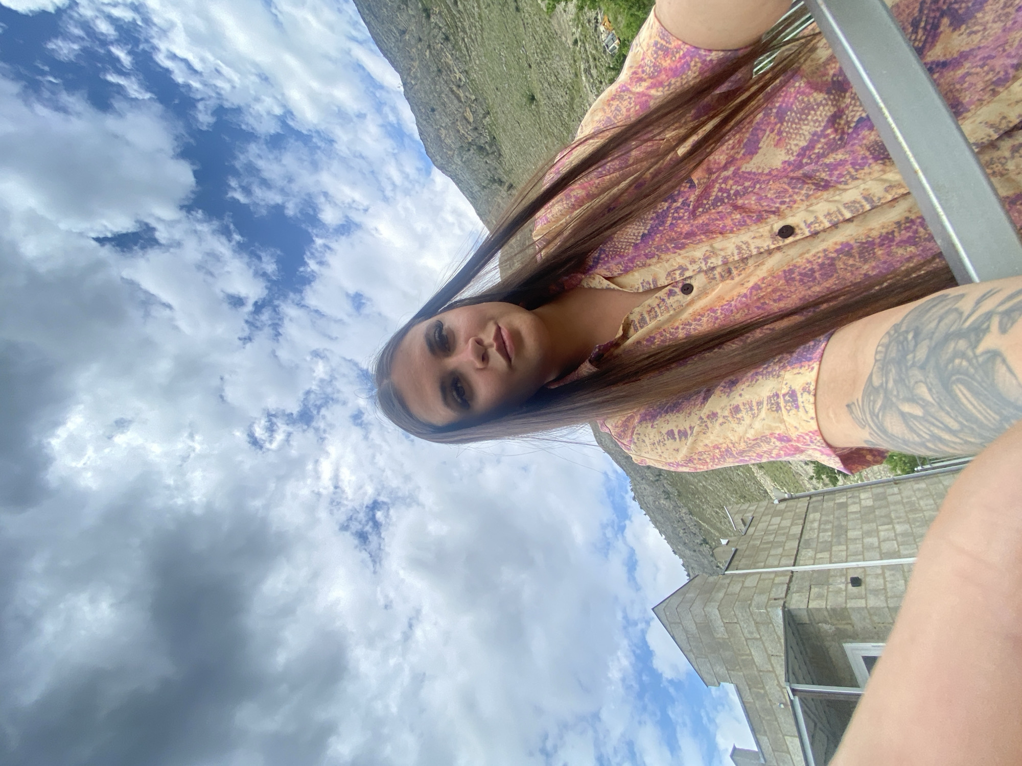 This is a low-angle selfie taken outdoors with a cloudy sky above. A woman with long brown hair, wearing a patterned button-up shirt, is the central subject. Her right arm is extended towards the camera, and a large tattoo is visible on her left forearm. In the background, a gray, unfinished brick building with a metal roof structure is situated on the left. Further in the background, a steep, barren mountain slopes upwards, partially covered in greenery. The location is identified as Chalda, Russia.