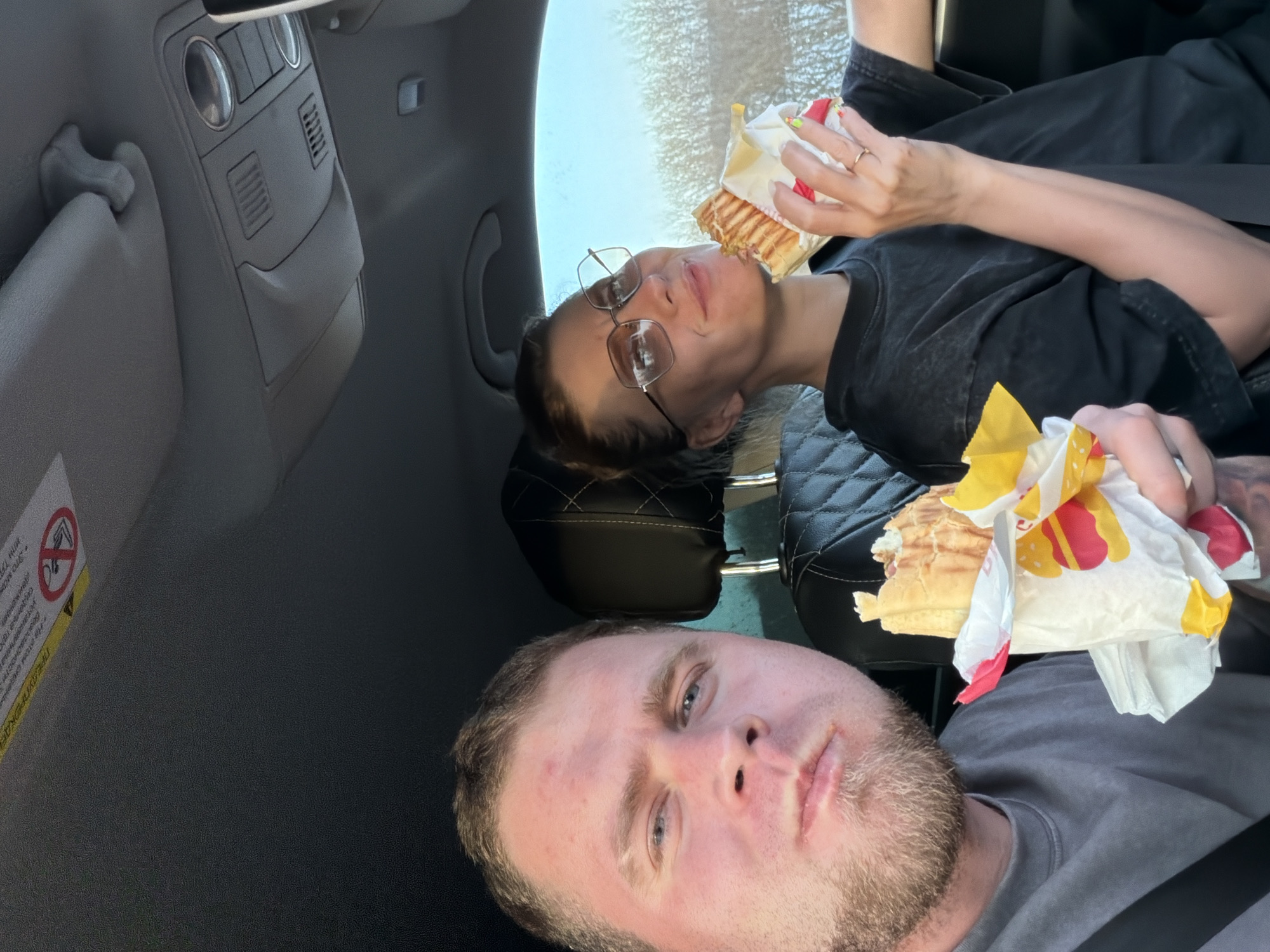 Two people are in the front seats of a car. A fair-skinned man with short blonde hair and a beard is in the driver's seat. A fair-skinned woman wearing glasses is in the passenger seat. Both are eating sandwiches, which are partially wrapped in paper packaging. The car interior is gray. There is a warning sticker on the sun visor. The specific location in the city of Chernovskoe, Russia cannot be determined from the provided image.