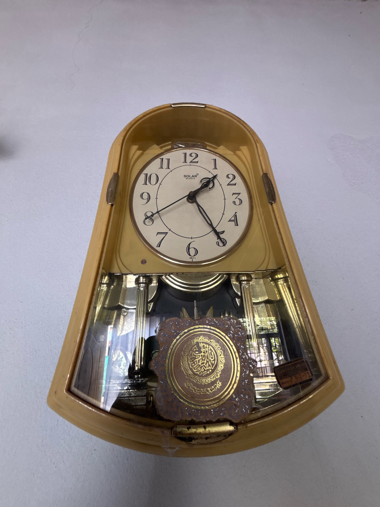 A pendulum wall clock with a light yellow casing hangs on a white, textured wall. The clock face is cream-colored with black Arabic numerals and a gold-colored inner rim. The hour hand points to approximately 8, and the minute hand points to approximately 50. The clock is branded "SOLAR QUARTZ". Below the clock face, a glass-covered compartment reveals a pendulum and decorative golden elements, including an ornate circular emblem with Arabic calligraphy. The overall impression is that this is a decorative clock, possibly with religious significance due to the calligraphy. The location is specified as Ghataprabha, India, though the image itself does not provide visual cues to confirm this specific location.