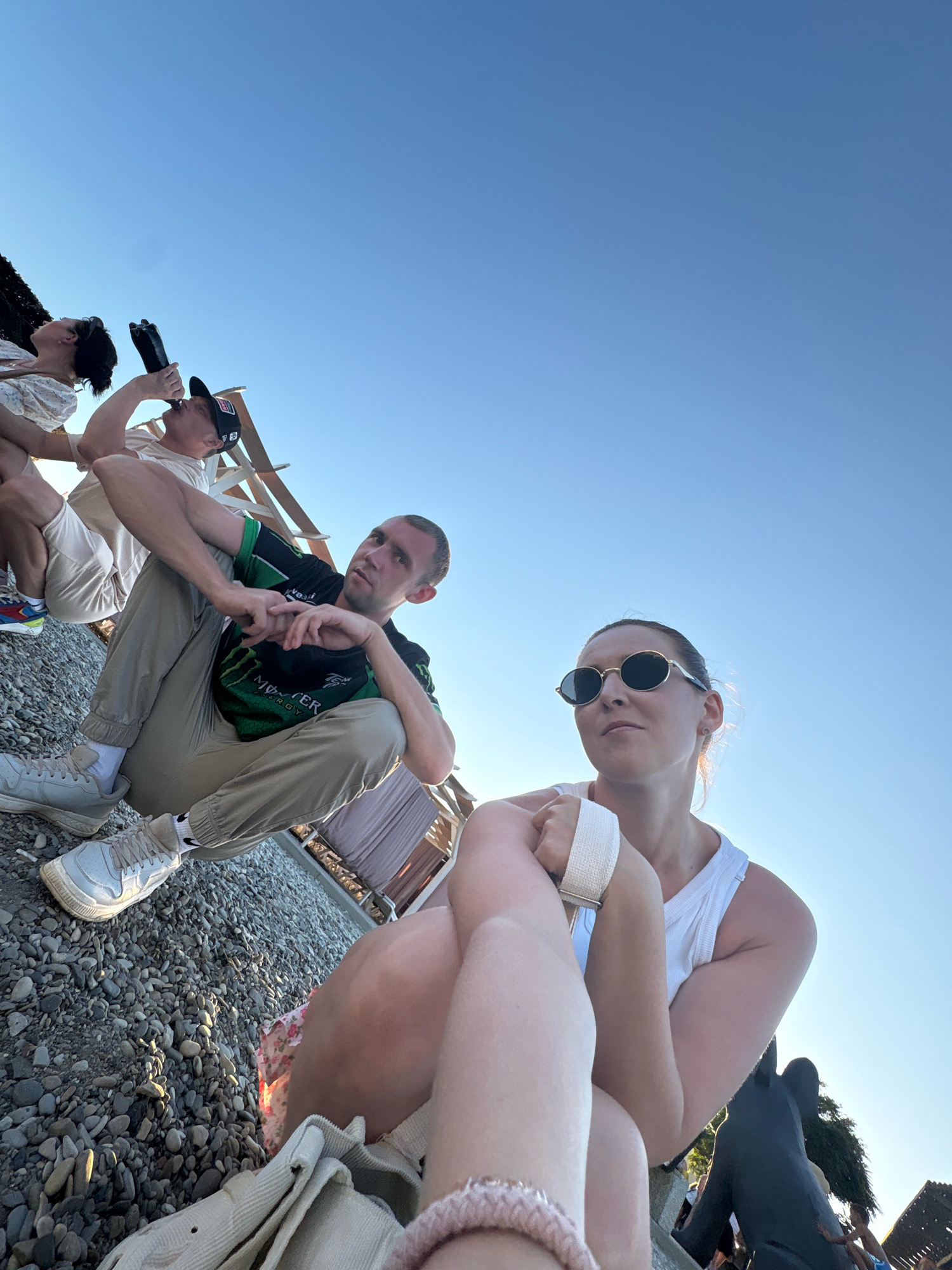 This is a wide-angle, low-angle selfie taken outdoors in Arkhipo-Osipovka, Russia, on a pebble beach, with a clear blue sky dominating the background. In the foreground, a person's legs and torso are visible, wearing a white tank top and a floral-patterned skirt. To the right, a woman with sunglasses and a white tank top is partially visible, looking off to the side. To the left, a man in a green and black t-shirt and khaki pants is sitting with his hands clasped. Behind him, another person is drinking from a bottle, wearing a light-colored t-shirt and a baseball cap. In the background, there are some beach structures and trees, suggesting a resort area. The lighting indicates it is daytime, likely late afternoon due to the warm light.