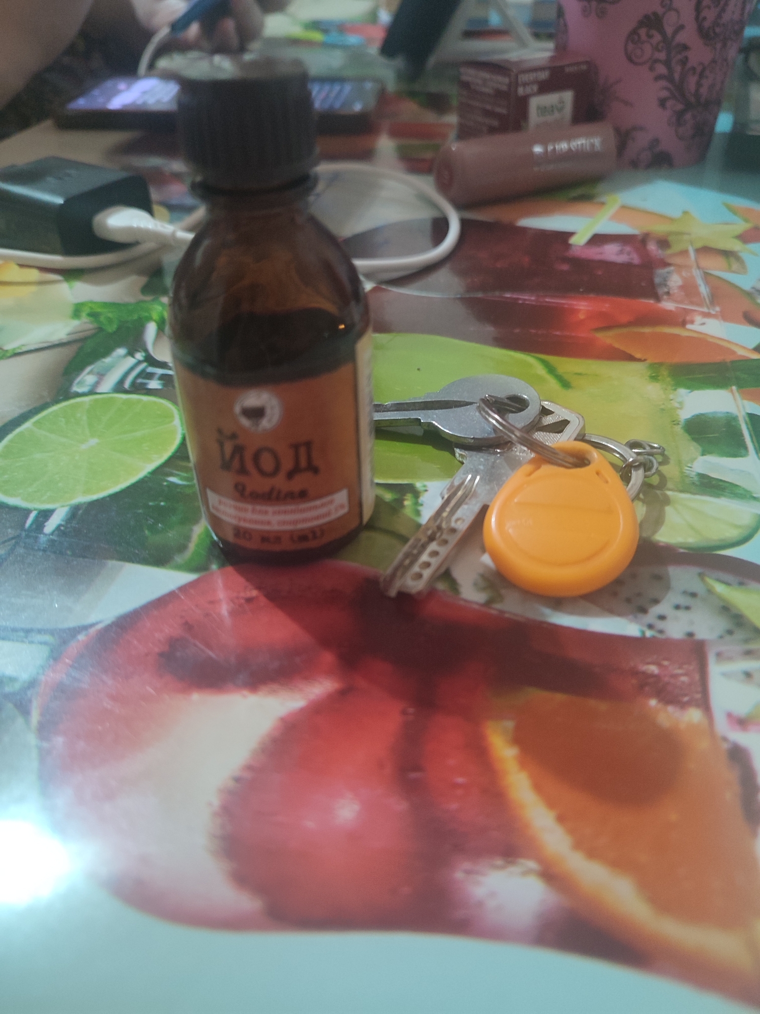 The image depicts a close-up of a table surface with various objects. In the foreground, a small brown glass bottle with a dark cap is visible, labeled in Cyrillic script with "Йод" (Iodine) and in Latin script with "Iodine." The bottle appears to contain a dark liquid. To the right of the bottle, a set of keys is present, attached to a keychain with an orange, teardrop-shaped fob. The table surface itself is decorated with a colorful pattern that resembles sliced fruits, including what appears to be lime and possibly citrus or berries. In the background, out of focus, are other items including a smartphone, a black power adapter plugged into a white cable, and a pink decorative object. The overall scene suggests a casual setting, possibly a home or a desk area. There is no discernible information within the image that would indicate its specific location in Chernivtsi, Ukraine.