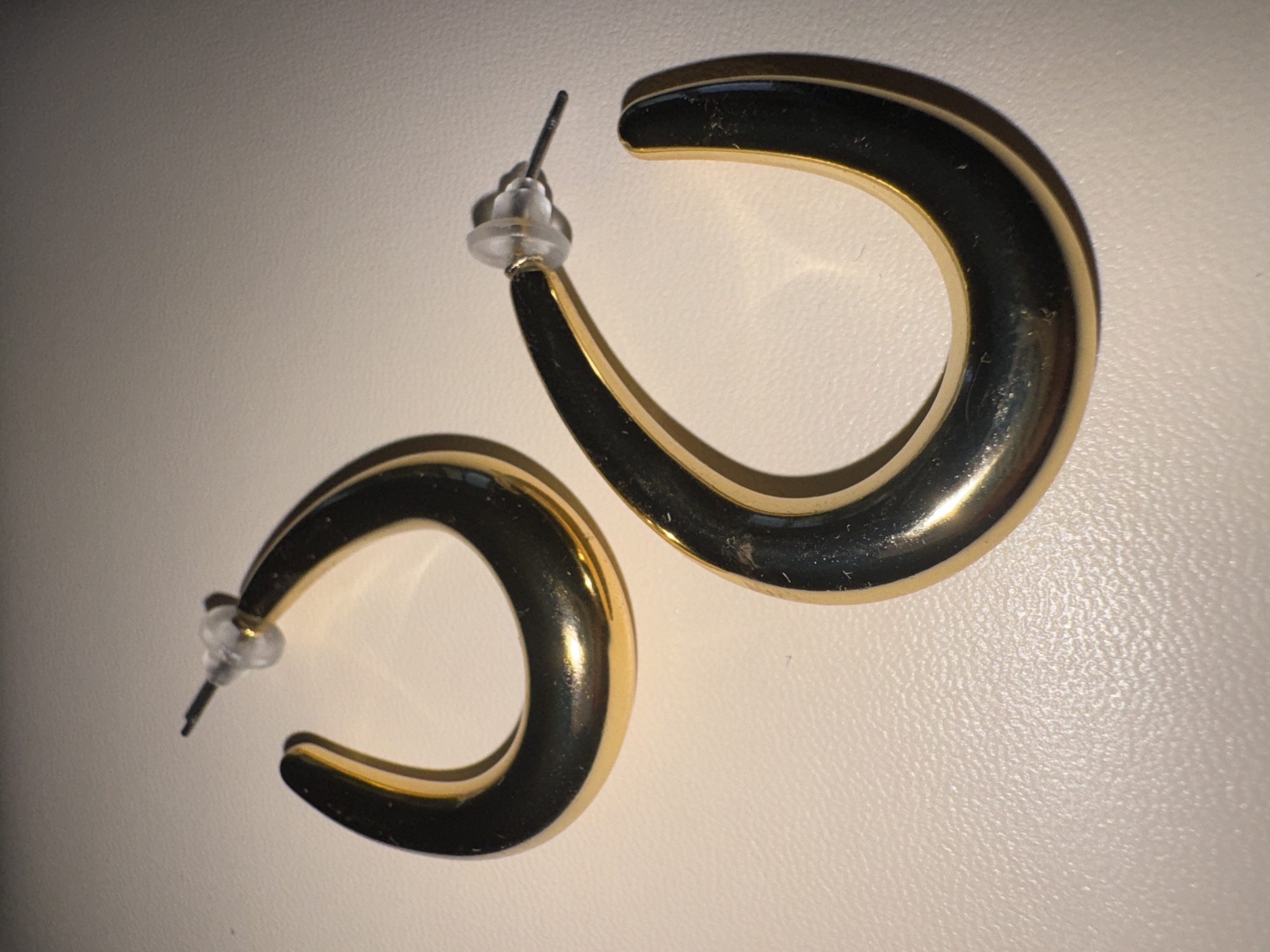 This image shows a pair of gold-colored hoop earrings against a textured, off-white background. The earrings are open at the bottom, forming a U-shape. One earring is larger and positioned to the right, while a smaller one is to the left. Both earrings have a polished, reflective surface, showing some minor scuffs and dust particles. The backs of the earrings have posts with clear plastic stoppers. The lighting suggests an indoor setting, likely a flat surface. There is no text visible in the image. The overall impression is a simple product shot of jewelry.
