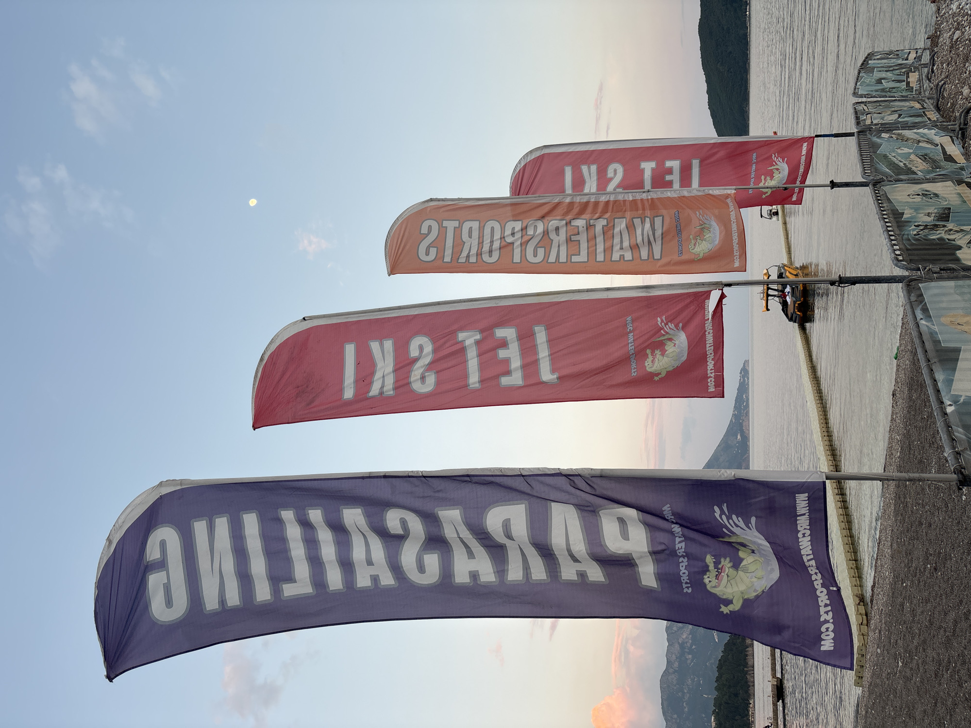 Here is a detailed description of the image:
The image shows a row of flags advertising water sports by the water's edge, likely in Budva, Montenegro. The flags are colorful and feature advertisements for parasailing and jet skiing. The flags are mounted on poles and are aligned along the edge of the water. In the background, there is a body of water, a coastline with mountains, and a pale sky with a hint of a moon. Adjacent to the flags on the right are a row of lounge chairs. A small yellow vehicle is visible further along the waterfront near the flags. The text on the flags is reflected due to the angle of the photo.