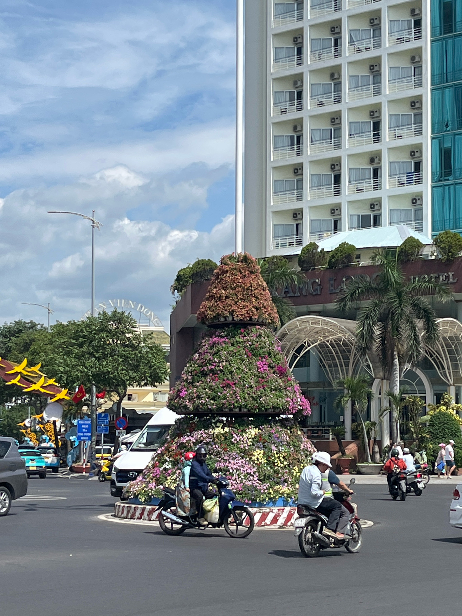 In Nha Trang, Vietnam, a roundabout is adorned with a large, tiered floral display shaped like a Christmas tree. The roundabout is situated on a sunny day with a partly cloudy sky. In the background, a tall white hotel with numerous balconies and air conditioning units dominates the skyline, with a turquoise glass building to its right. The hotel's signage, partially visible, reads "XUAN DONG HOTEL". Several people are present, with some riding motorcycles and scooters through the intersection. One man on a scooter wears a blue shirt and helmet, while another, further along, wears a white hat and a light blue shirt. Other individuals are visible in the distance, some walking. Traffic includes a white van, a blue taxi, and a dark gray car. Yellow decorative banners are strung across poles, and various flags are also visible. The roundabout itself features a white and red striped border around the floral arrangement.
